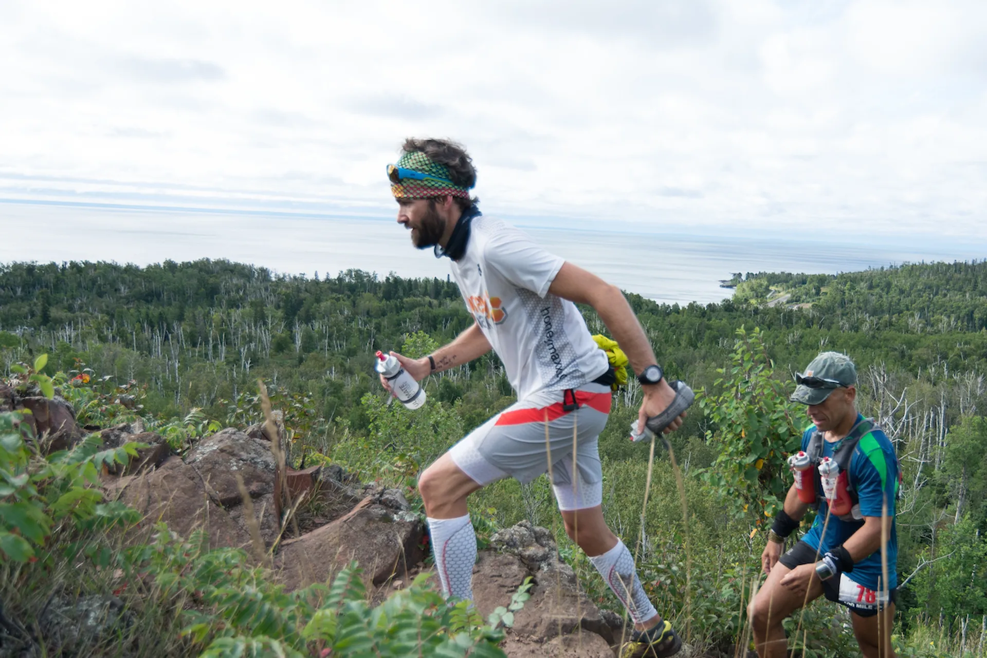 The image depicts three individuals engaged in a trail running event. The foreground shows a