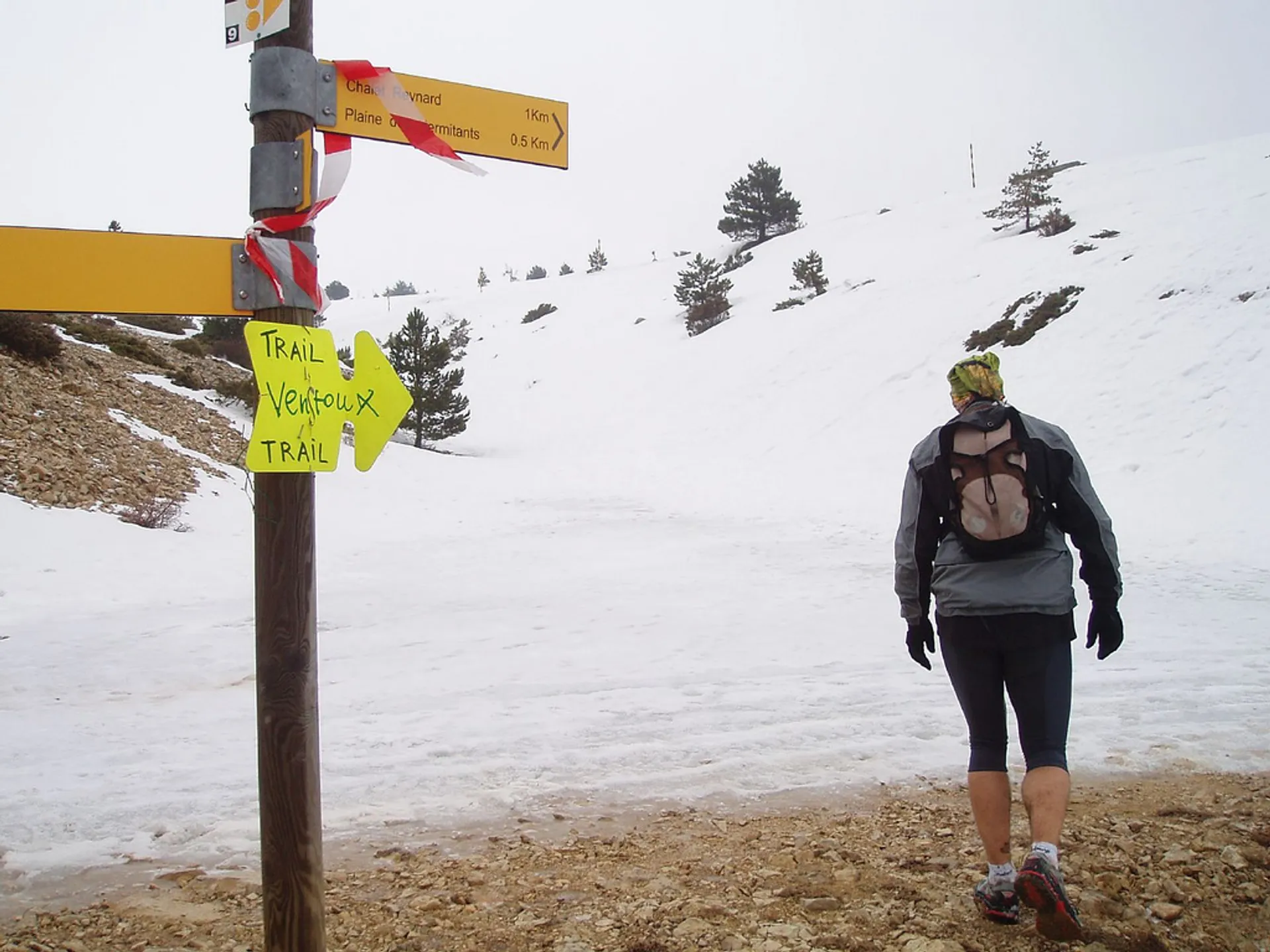 The image shows a person standing at a trail junction in a snowy mountain area.