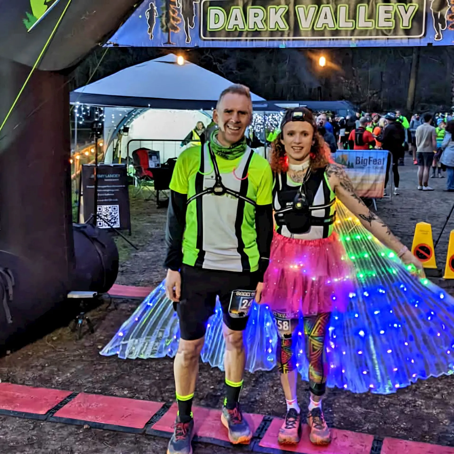 This image shows two people dressed in running gear, posing under a banner that says "Dark Valley." The person on the left is wearing a green and black athletic outfit. The person on the right is wearing a colorful outfit with illuminated wings. The setting appears to be an outdoor event or race.