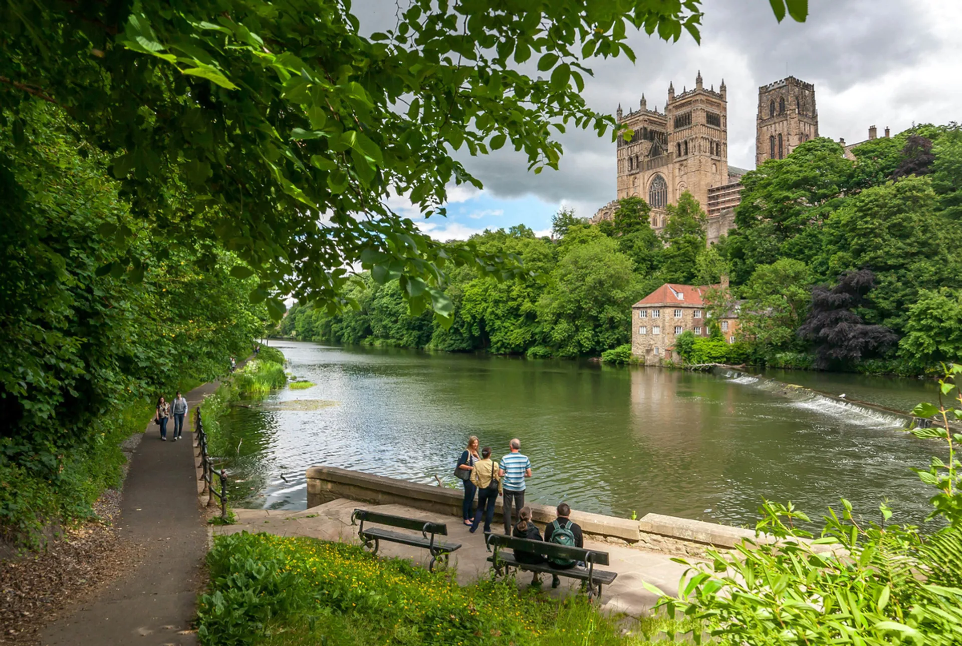 In the image, there is a picturesque outdoor scene featuring a river with calm waters. Alongside the river, there is a well-maintained footpath where a couple of people are walking and enjoying the view. Closer to the foreground, there are two individuals seated on a bench, seemingly in conversation or simply taking in the serene environment.

To the right of the riverbank, there are dense trees and shrubs contributing to the lush greenery of the area. Furthermore, in the background, towering above the trees, there is an impressive building with Gothic architecture, including large windows and two prominent towers. This building appears to be a cathedral or an abbey, giving the scene a sense of historical significance.

The overall ambience of the photo