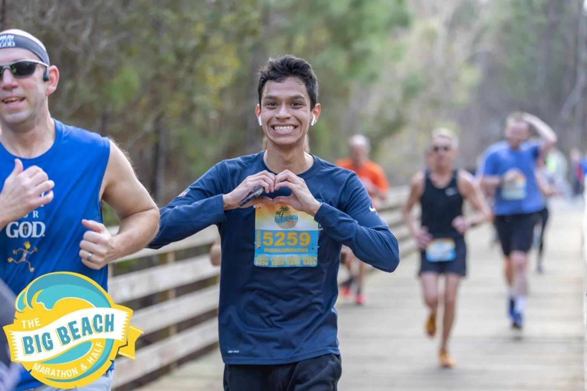 The image shows people participating in a running event called "The Big Beach Marathon & Half." One runner in the foreground is making a heart gesture with his hands and appears to be smiling. Other runners are visible in the background on a wooden path surrounded by trees.