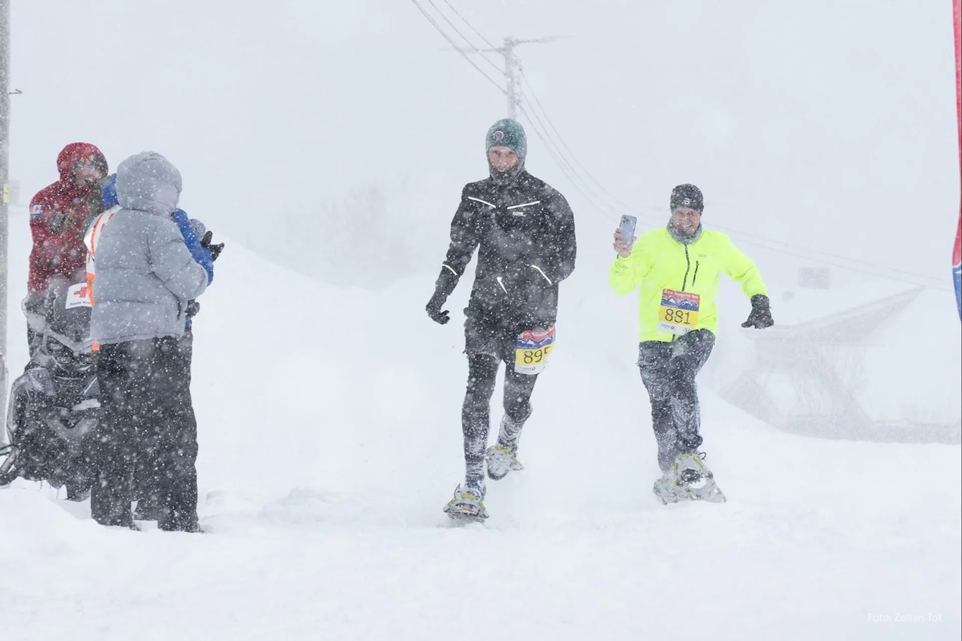 The image depicts a snowy scene where a group of people are engaged in a winter activity, likely a competitive event given the presence of bib numbers on the participants. Two of the individuals in the foreground appear to be running or racing while wearing snowshoes, which is a specialized winter sport. It's snowing heavily, as indicated by the white snowflakes visible against the participants and the reduced visibility in the background. There are also onlookers or officials standing to the side, one of whom seems to be holding some event-related equipment, possibly for timing or for assistance. The participants and onlookers are dressed in winter gear to stay warm, including jackets, gloves, and hats, indicating cold conditions. The presence of overhead cables suggests this