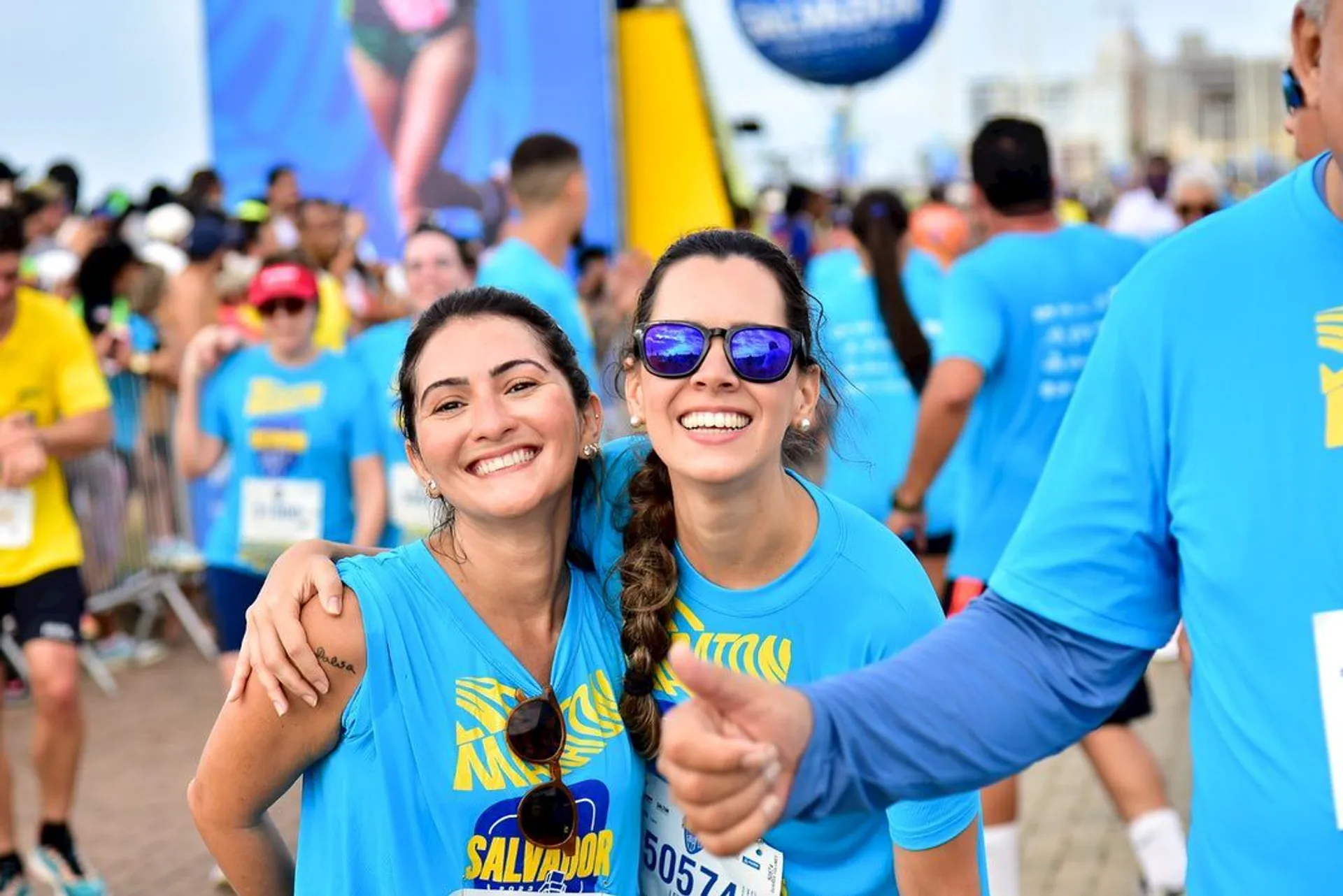 The image shows two women posing with their arms around each other's shoulders, smiling brightly at the camera. They are wearing matching blue event t-shirts, suggesting they are participating in a group activity such as a run or a public event. The woman on the left has her hair tied back, and the woman on the right is wearing sunglasses and also has her hair tied back in a braid. They both appear to be very happy and enjoying themselves.

In the background, there is a crowd of people who also seem to be wearing similar shirts, indicative of a large gathering or public event such as a marathon, charity run, or outdoor festival. There are event structures like start/finish line arches and banners, adding to the festive atmosphere. The