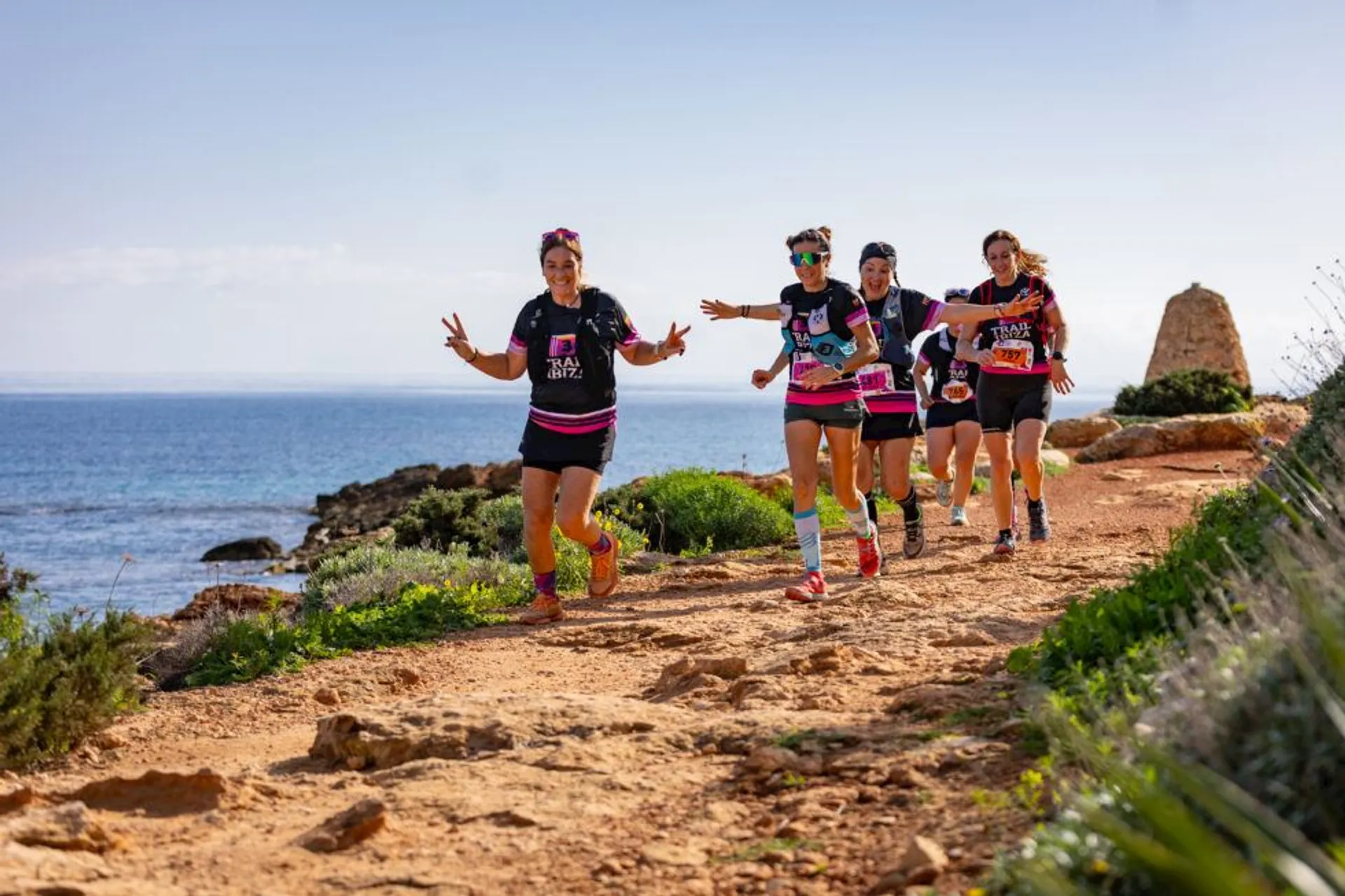 The image shows a group of people running on a scenic coastal trail. They seem to be participating in a running event, as indicated by their numbers and matching shirts. The background features a clear sky and ocean view, creating a vibrant outdoor setting.