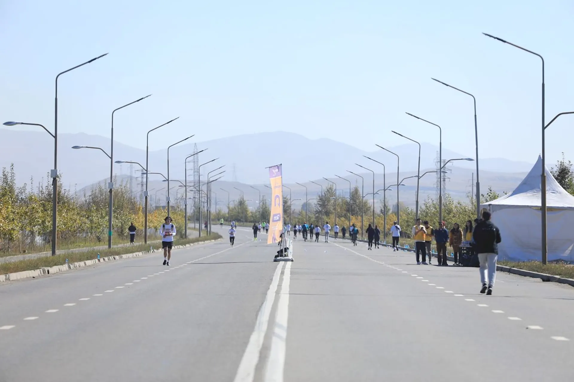 The image shows a road with people running and walking, likely participating in a marathon or running event. There are streetlights lining the road and a few trees visible on the sides. Far in the background, there are mountains under a clear sky. The road is clear of cars, suggesting it's been closed for the event.