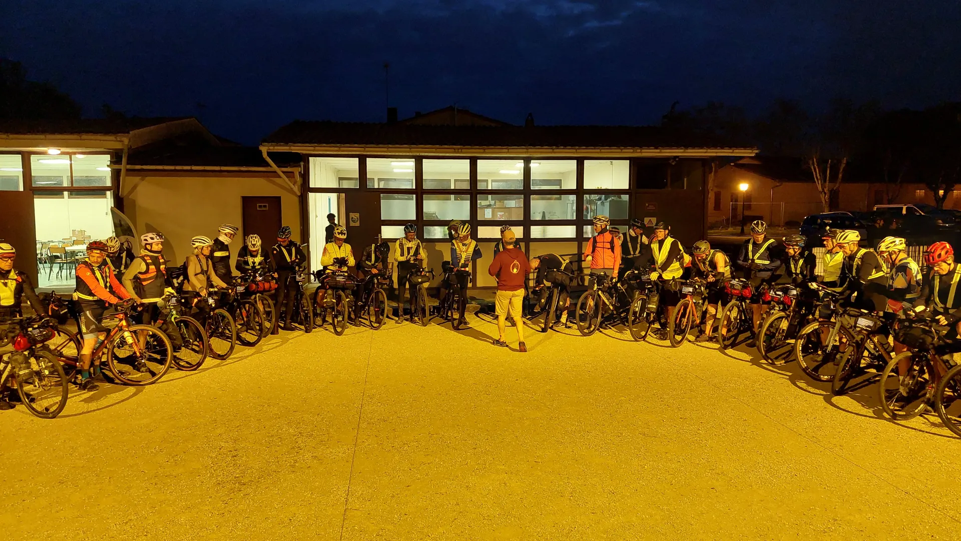 The image shows a group of cyclists gathered together, forming a semi-circle around someone standing in front of them. It appears to be taken outdoors near a building in the evening or early morning, as the sky is dark. The cyclists have gear and their bikes with them, suggesting they are preparing for or concluding a ride.