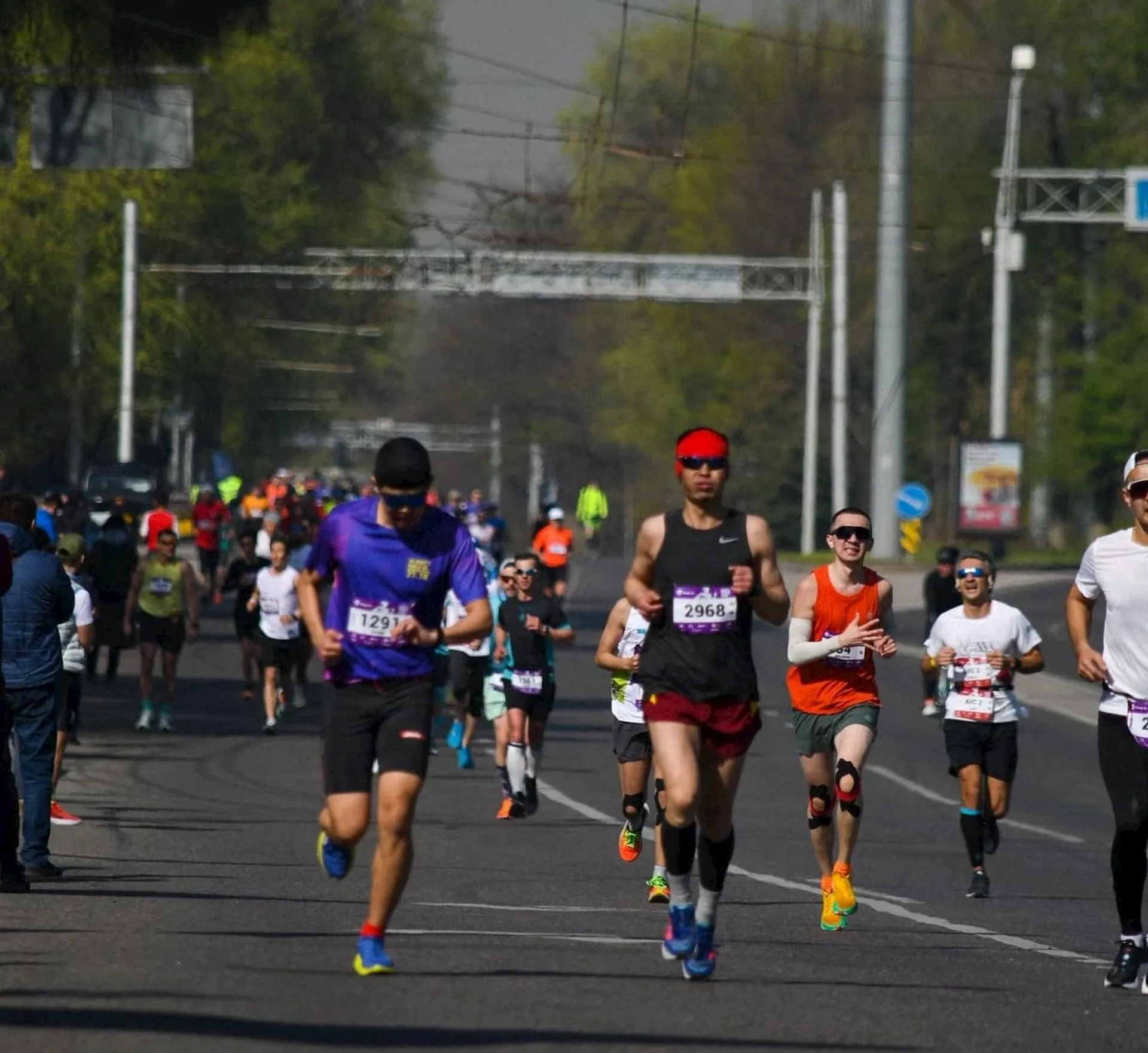 The image shows a group of people participating in a road race or marathon. They are running on a street, and some are wearing race bibs with numbers. There are trees lining the road and other participants visible in the background.