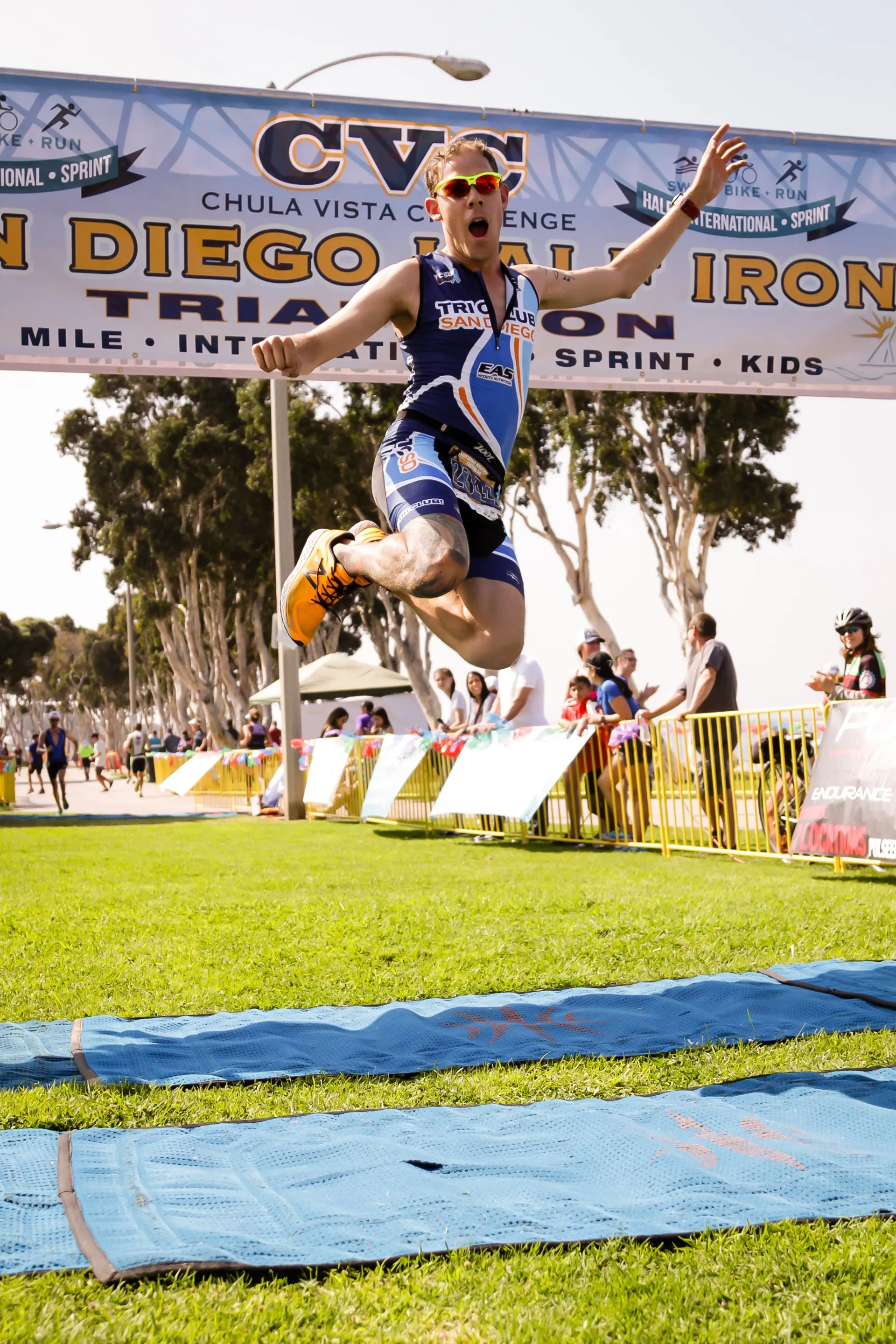 The image depicts an athlete crossing the finish line at what appears to be a triathlon event, given the "SAN DIEGO INTERNATIONAL TRIATHLON" banner overhead. The athlete is wearing sports attire, a race number, and sunglasses, and is captured in an expressive pose with arms raised and mouth open, likely cheering or celebrating the moment of victory. The background is an outdoor setting with trees and grass, indicative of a park or similar open area. Various event paraphernalia, such as fencing, banners, and a timing mat, are also visible, typically used for organizing and timing the race.