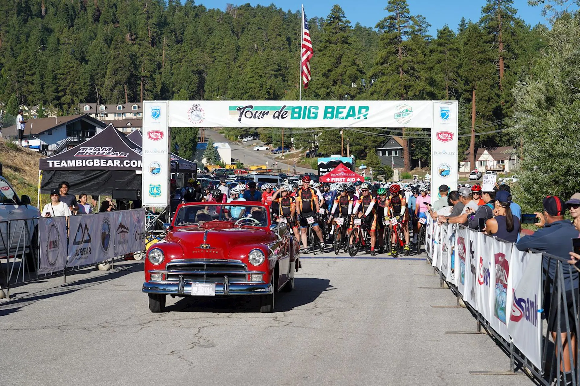 The image shows a starting line for a cycling event called "Tour de Big Bear." In the foreground, there's a classic red car leading the cyclists, who are lined up behind it. There are spectators on either side behind barricades, and the event is taking place in an outdoor area surrounded by trees and hills. Banners and flags are displayed around the area.
