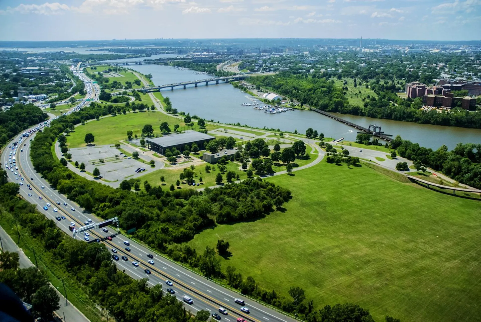 This image shows an aerial view of a cityscape with a river running through it. There are bridges crossing the river and a significant amount of green space and trees. Roads with moving traffic can be seen along with some buildings. The overall scene appears to be a mixture of urban and natural landscapes.