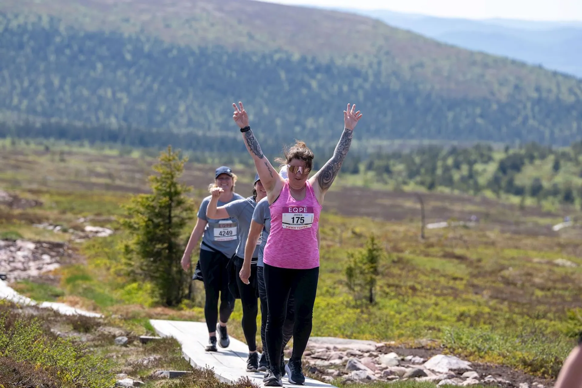 The image shows three people walking or running on a pathway in a hilly or mountainous area with greenery. The person in front is wearing a pink tank top, has their arms raised, and appears to be participating in an event, as indicated by the number bib they are wearing. The scene suggests an outdoor race or marathon.