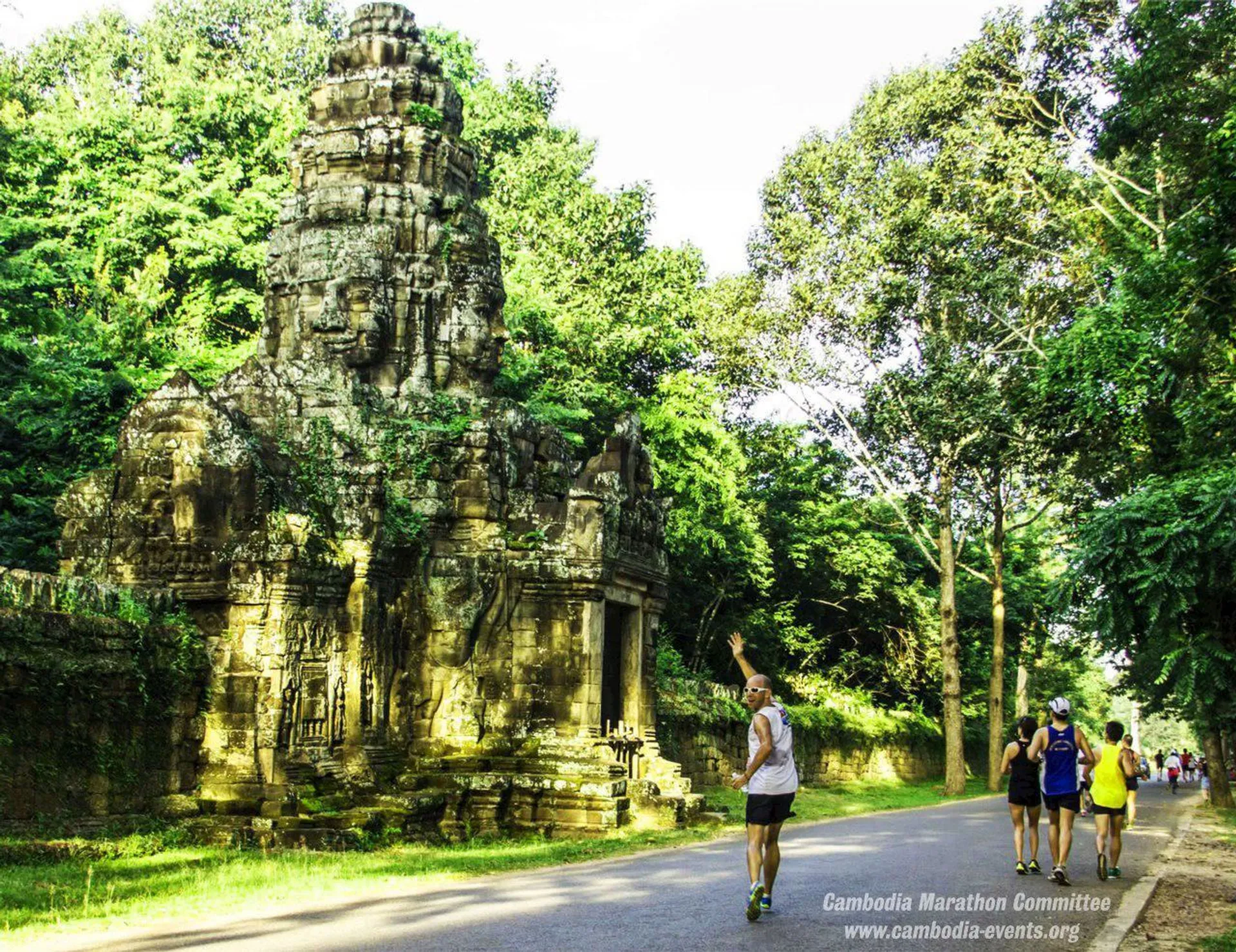 This image shows a group of people who appear to be jogging or running on a path flanked by lush green trees. On the left side of the path is an ancient stone tower with intricate carvings, suggesting that this location might be a historical site or ruins, possibly within a region rich in cultural heritage such as Southeast Asia. The watermarks indicate that the photo might be related to a marathon event organized by the Cambodia Marathon Committee, thus the location could be somewhere in Cambodia, famous for its historic temple complexes like Angkor Wat. Overall, the combination of active individuals and the serene, historic backdrop suggests an event that marries fitness with cultural exploration.