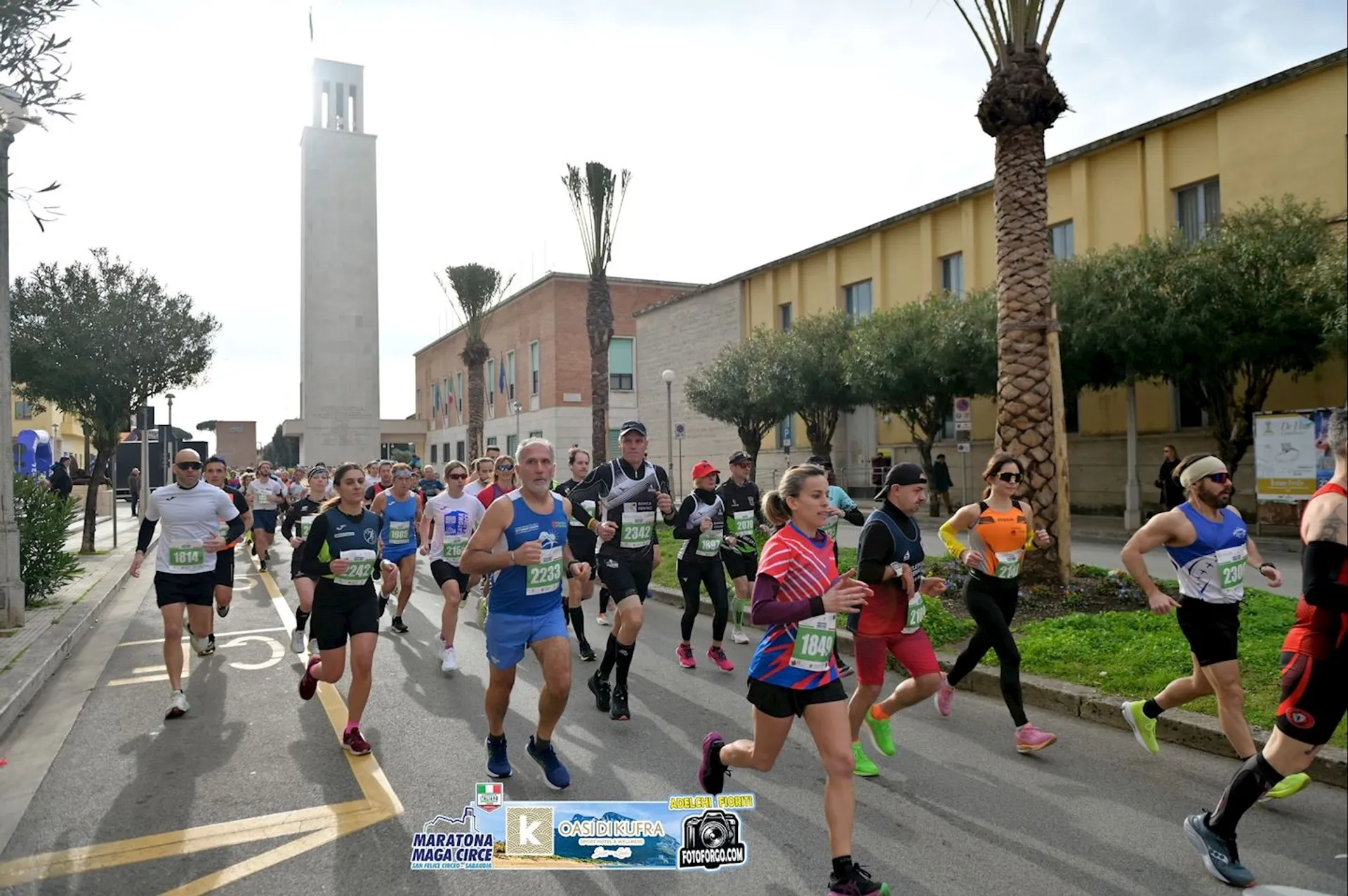 The image shows a group of runners participating in a race on a street. They are wearing race bibs and athletic gear. There are buildings and trees lining the street, and a prominent tower in the background. Logos related to the event are visible at the bottom of the image.