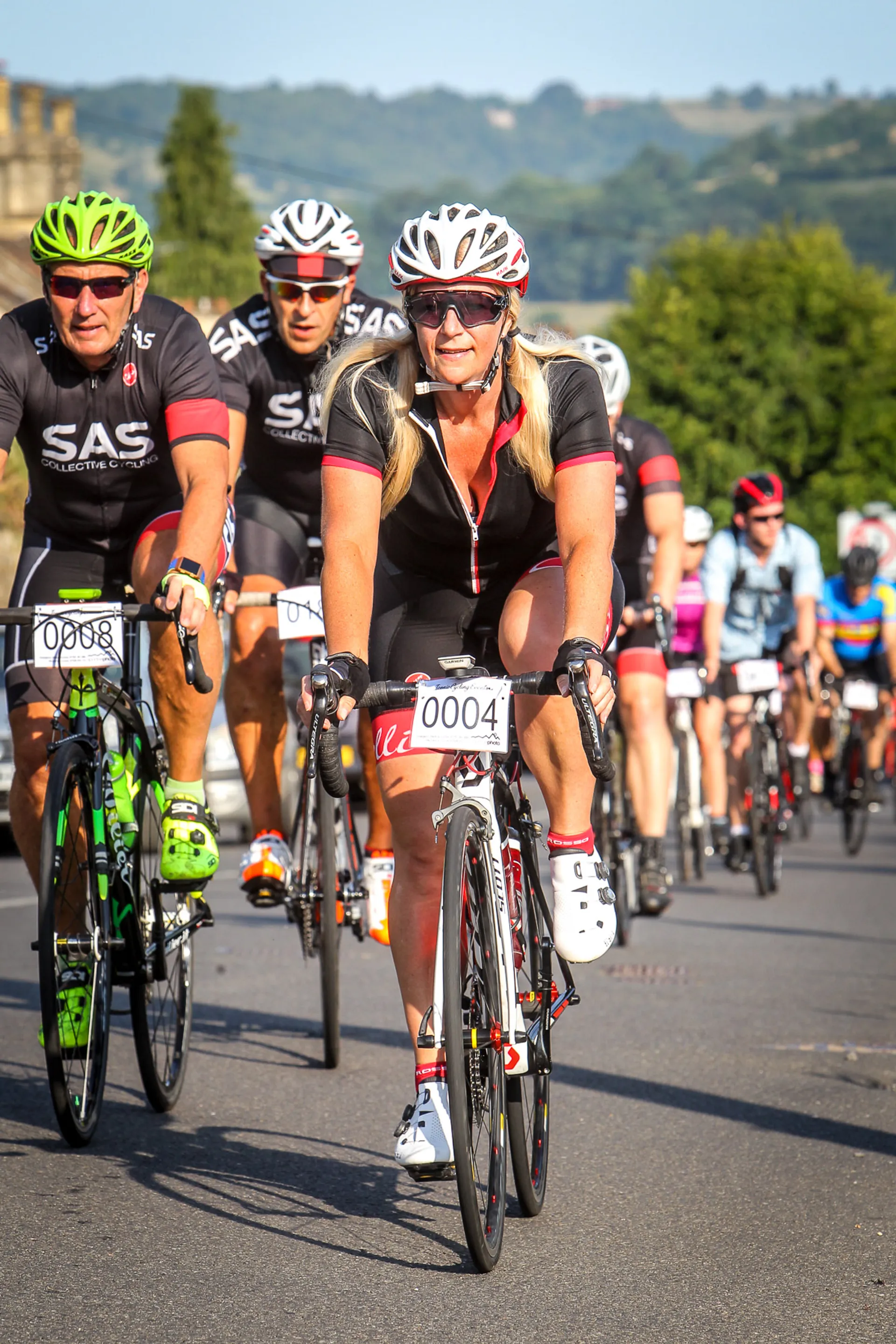 The image displays a group of cyclists participating in a bike race or organized cycling event. At the forefront, there's a woman wearing sunglasses, a helmet, and a cycling outfit with a visible race number "0004." She's riding a road bike with drop handlebars, which suggests this could be a road cycling event. Other cyclists are visible in the background, indicating that this is a well-attended event. Everyone appears to be wearing proper safety gear, such as helmets, and focused on their riding. The scenery includes a backdrop of gentle hills, indicating the event is taking place in a region with such geographical features. The weather appears sunny and favorable for an outdoor cycling event.