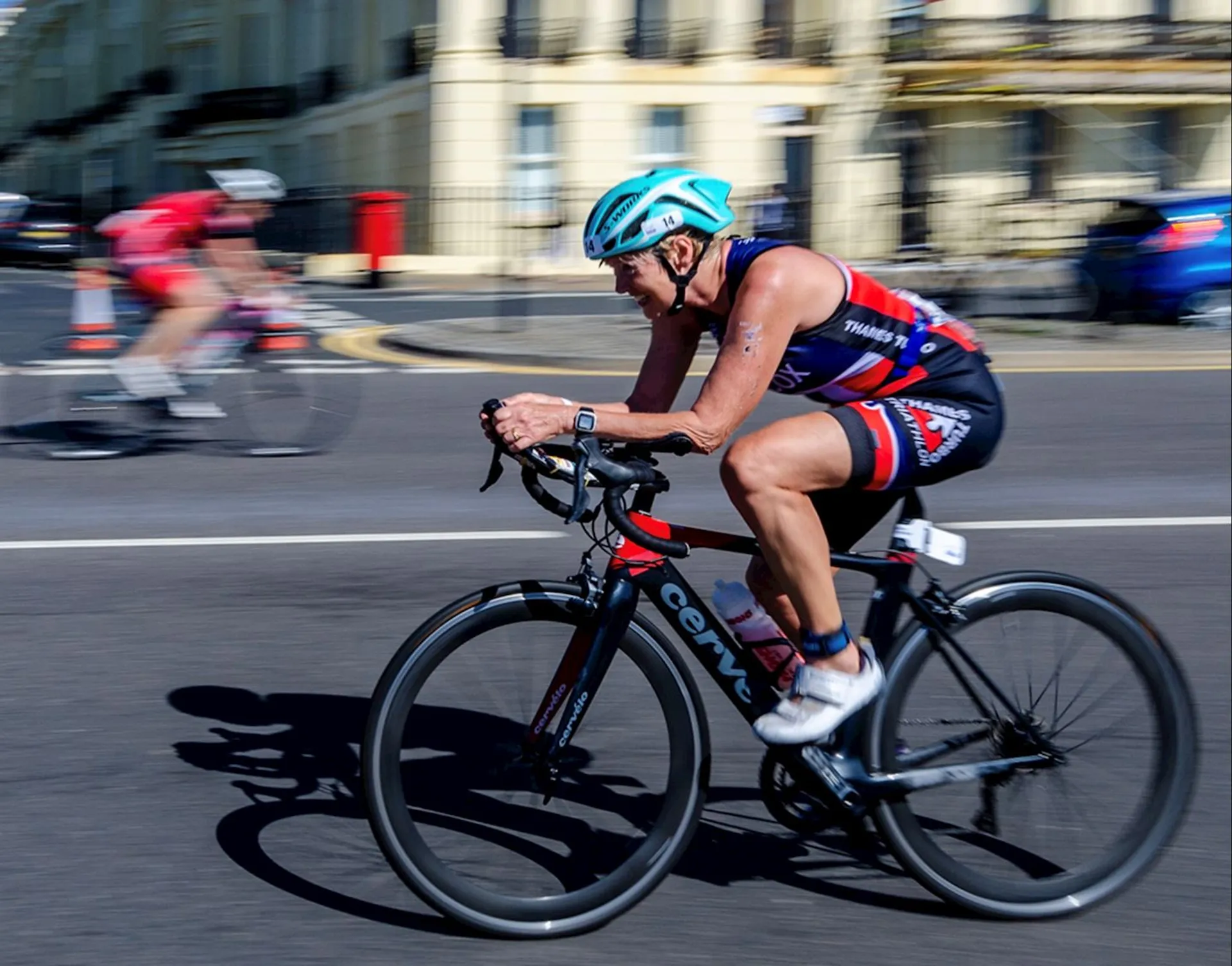 The image shows a cyclist in motion on the street, wearing a blue helmet, sunglasses, and a cycling outfit. The background is blurred, indicating speed, and there's another cyclist in the background.