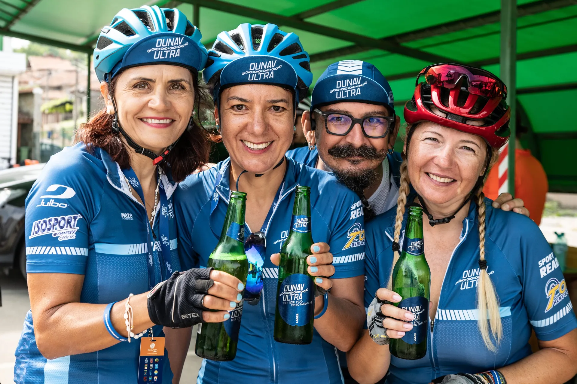 The image shows a group of four smiling cyclists, three women and one man, wearing matching blue and white cycling outfits with the name "DUNAI. Ultra" on them. They appear to be in a celebratory or social setting, each holding a green bottle that could normally be associated with beer or a similar beverage. They are also wearing cycling helmets, suggesting they either just finished a ride or are taking a break. They seem to be enjoying each other's company in an outdoor setting, possibly at a cycling event or a gathering related to cycling. The presence of tents or shelters in the background indicates they might be at a structured event with facilities for participants.