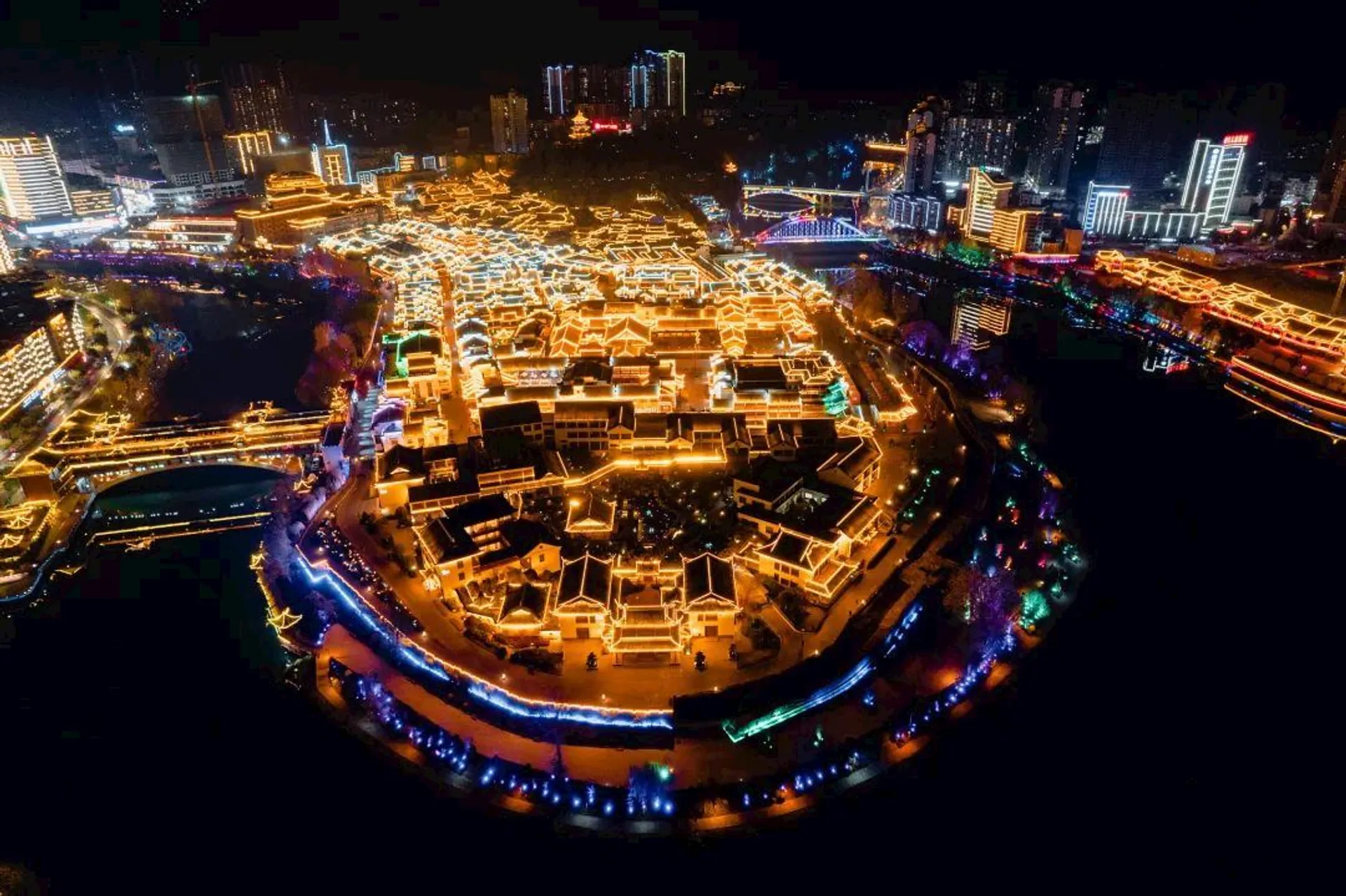 This image shows a vibrant nighttime cityscape with a focus on a cultural area, possibly featuring traditional-style buildings illuminated with golden lights. The scene includes winding pathways or streets surrounded by water and modern city buildings in the background also adorned with colorful lights. The combination of traditional architecture and modern elements creates a striking contrast.