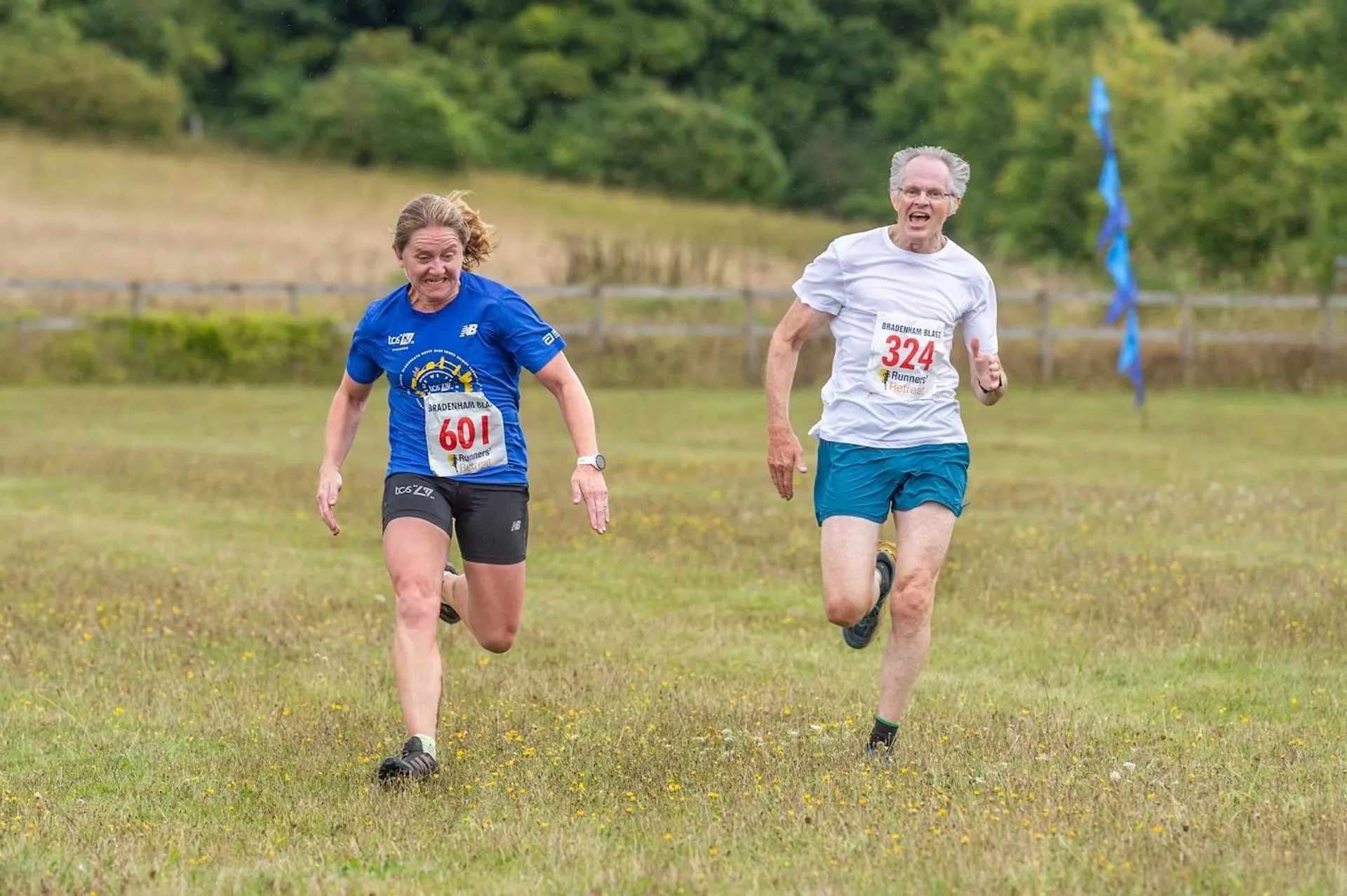 The image shows two people running in a grassy field, likely participating in a race. Both are wearing shirts with numbers attached. The area is open and appears to be part of an outdoor event, with trees and some flags or markers visible in the background.
