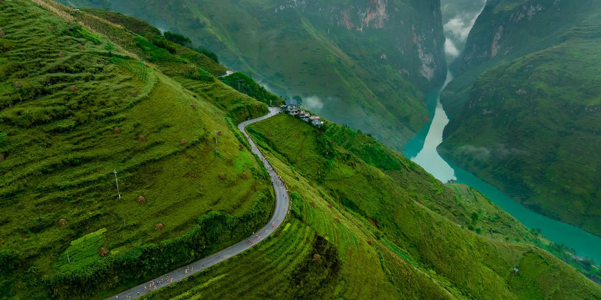 This image shows a stunning landscape featuring a winding road cutting through lush green hills. The hills are terraced, and in the background, there's a deep, narrow valley with a river flowing through it. The scenery is vibrant and serene, likely depicting a rural or mountainous region.