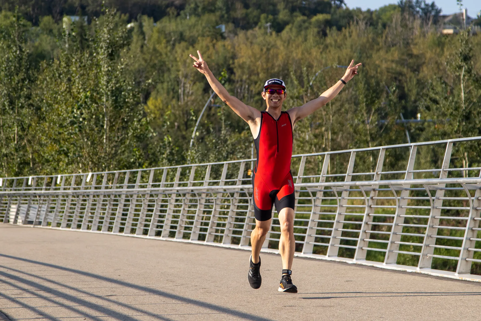 The image shows a person running on a paved path with a metal railing. They are wearing a red athletic outfit and sunglasses and have their arms raised in a celebratory gesture. The background features trees and a clear sky.