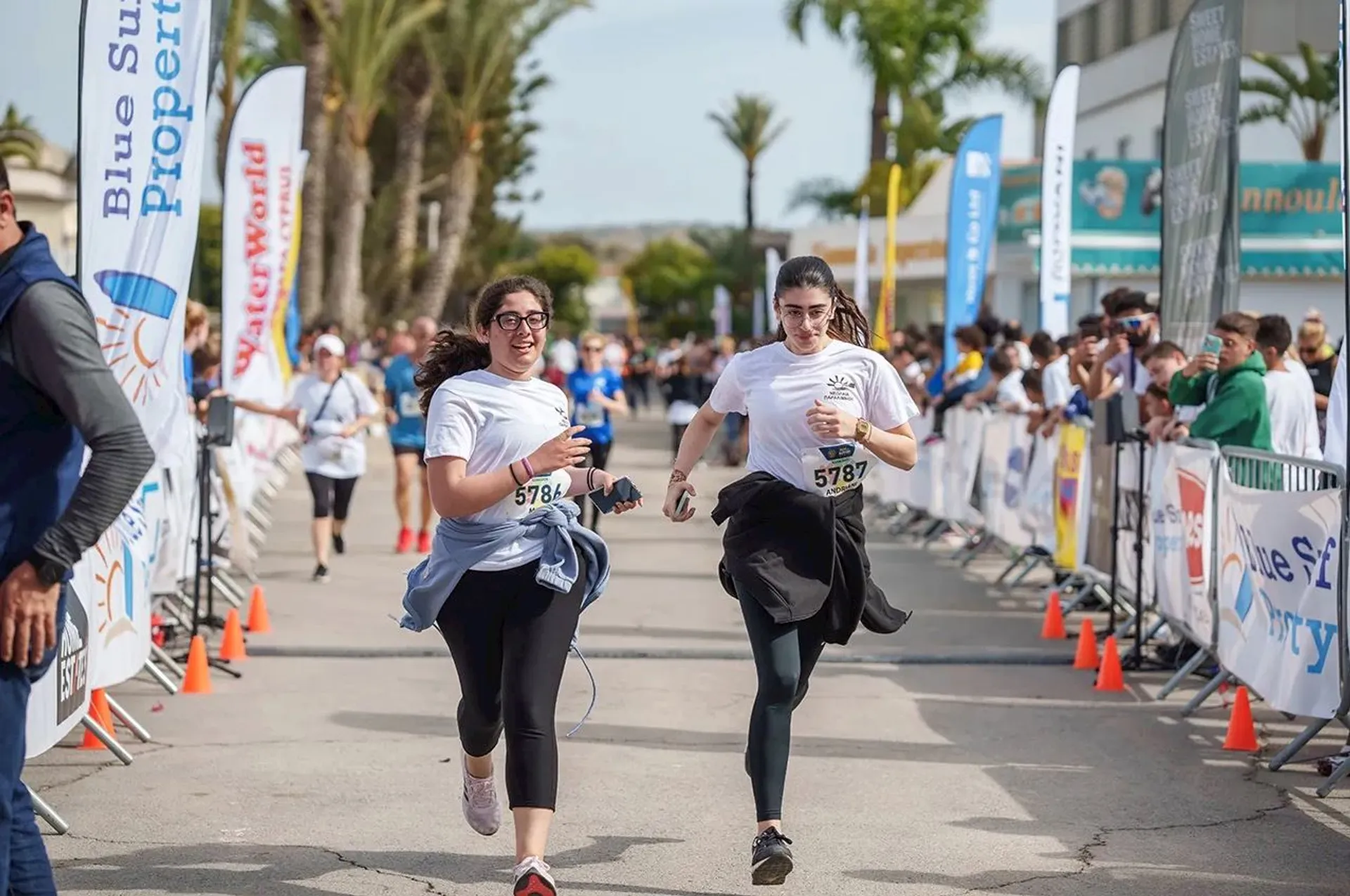 The image shows two people running in a race. They are wearing race bibs and are on a road with spectators on the sides. There are banners and flags along the route, suggesting it's an organized event. The background is lined with palm trees and some buildings.