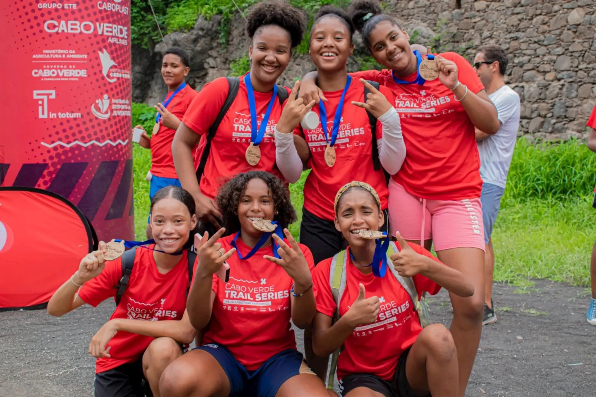 The image shows a group of six people wearing red shirts with "Cabo Verde Trail Series" written on them. They are posing for the camera, holding up medals and smiling. They appear to be part of a sporting event, likely related to running or trail series activities.