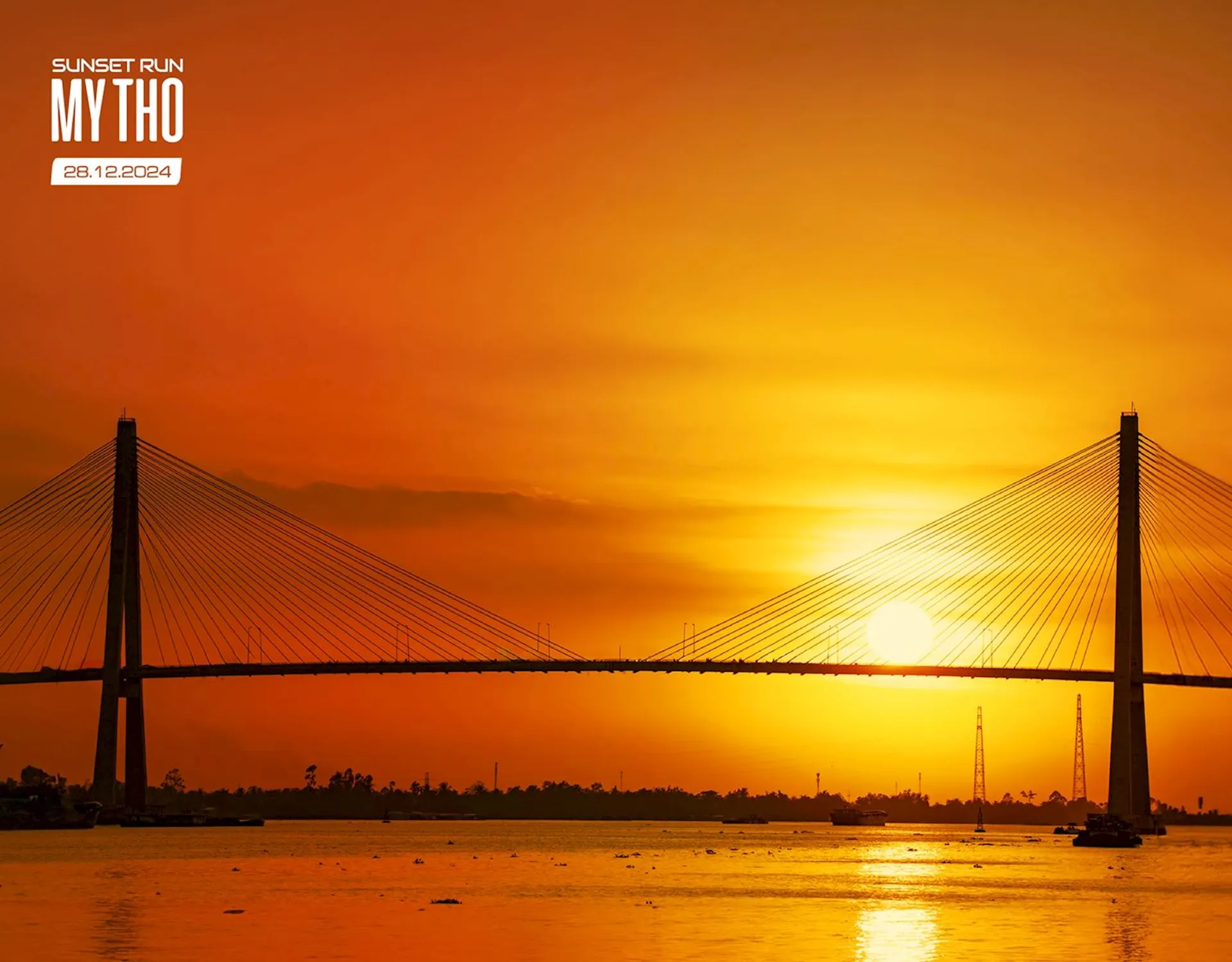This image features a beautiful sunset scene with the sun low in the sky, casting a warm, golden hue over the scene. A large cable-stayed bridge stretches across the frame, its silhouette prominent against the light of the setting sun. The body of water beneath the bridge reflects the sunlight, creating a shimmering effect. The sky is mostly clear with a few wispy clouds, enhancing the vibrant colors of the sunset. It appears to be a promotional image for an event called "Sunset Run My Tho" on the date given (presumably when the photo was taken or the event occurred), with the text in the upper right-hand corner overlaying the sky. My Tho may refer to a location, likely where this picturesque scene is set