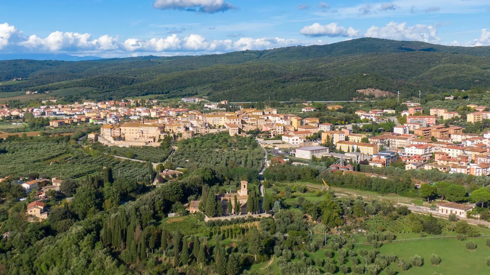 The image depicts an aerial view of a scenic landscape featuring a small town surrounded by lush greenery and hills. The town has a mix of buildings with red-tiled roofs and roads, nestled within trees and fields. In the background, there are forested hills extending towards the horizon under a partly cloudy sky.