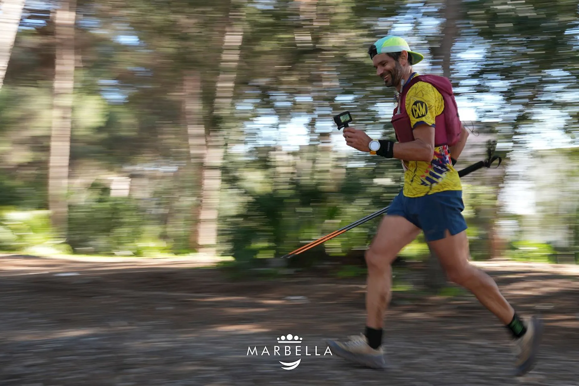 The image shows a person running through a forested area. The runner is dressed in athletic gear, including shorts, a shirt, a cap, and is holding what appears to be a smartphone in one hand. The background appears blurred, indicating motion. The runner also has a hydration pack and trekking poles. The word "Marbella" is displayed at the bottom of the image.