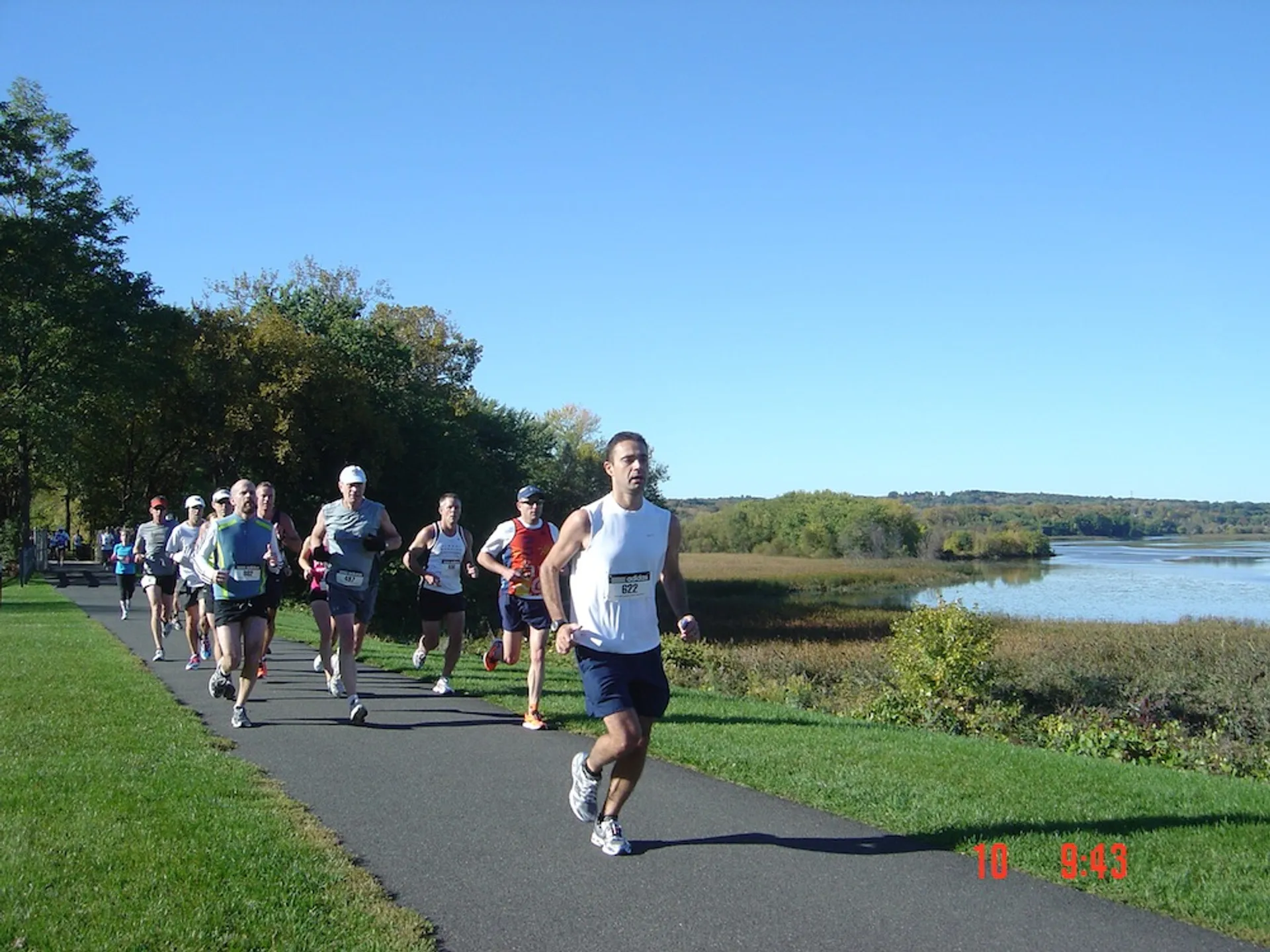 The image shows a group of people running on a paved path alongside a body of water. The setting appears to be outdoors in a park or natural area, with trees and grassy surroundings. The runners are wearing athletic gear, and it seems to be a sunny day.