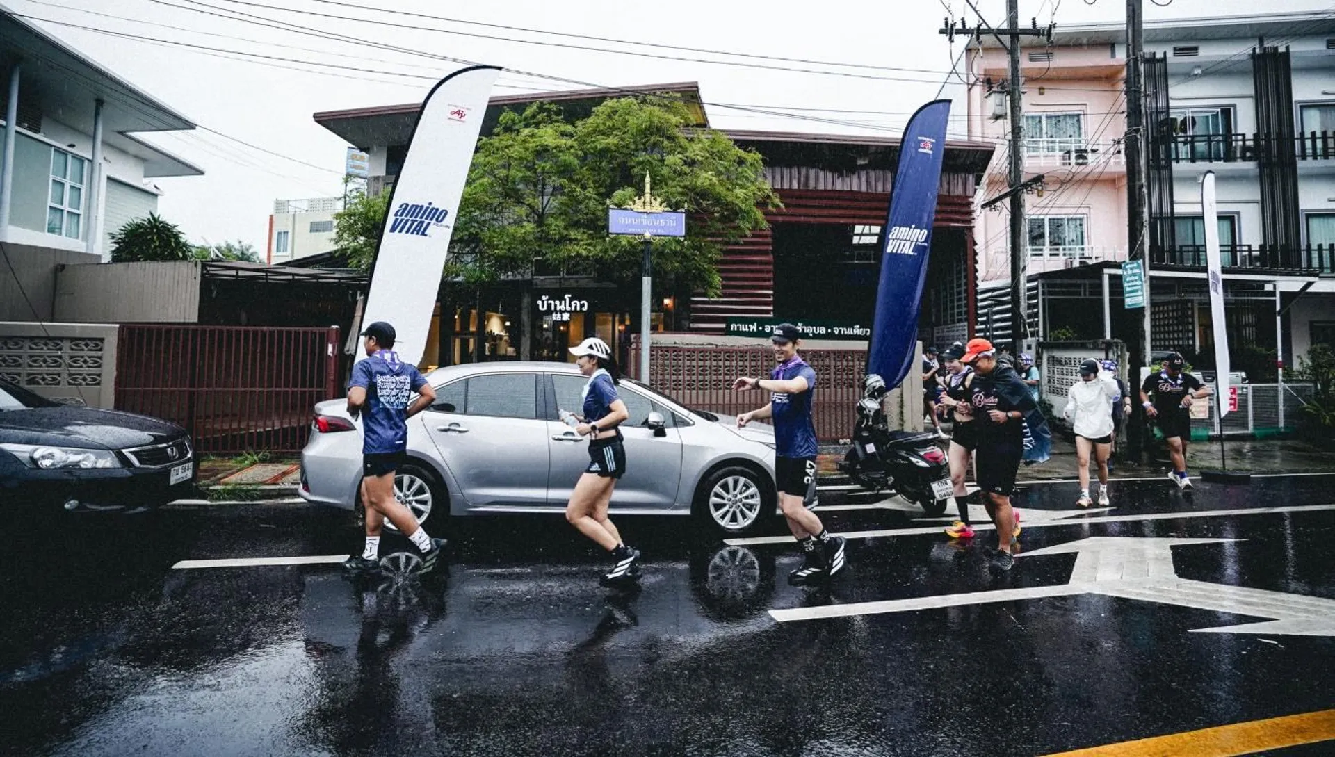 The image shows a group of people running on a wet street. They are wearing athletic clothing, and some have hats. There are two banners by the side of the street, and cars are parked nearby. The background features buildings and trees. It appears to be a running event or gathering.