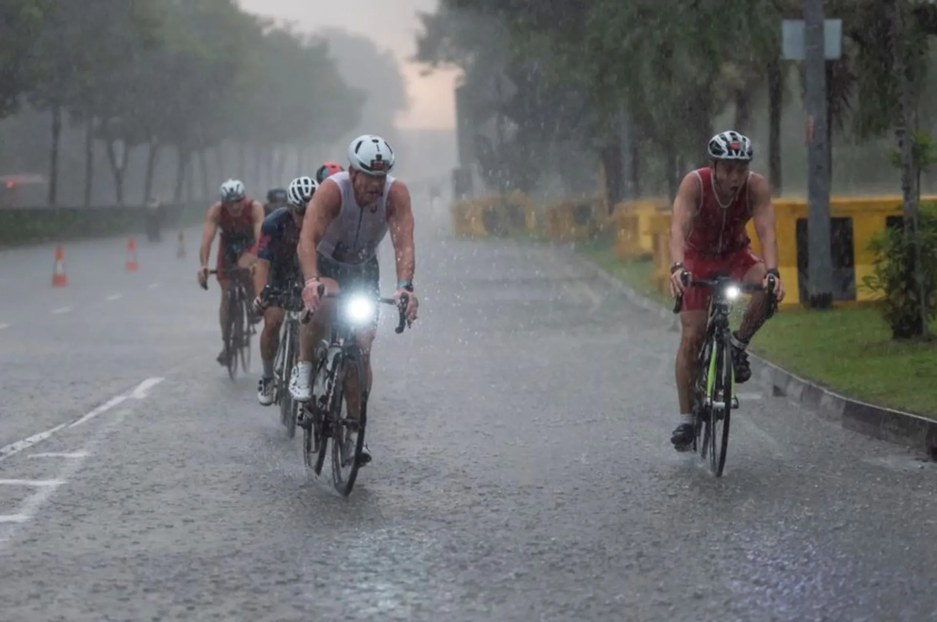 The image shows a group of cyclists riding their bikes on a wet road, presumably during a heavy rain shower. The cyclists are equipped with race numbers, indicating that they are participating in a competitive event, likely a triathlon or a cycling race. They are all wearing helmets for safety, and the one in the foreground has a light on their bike, which helps with visibility in the rain. The trees and overcast sky suggest that the weather conditions are quite challenging for the event, but the athletes are pressing on despite the inclement weather.