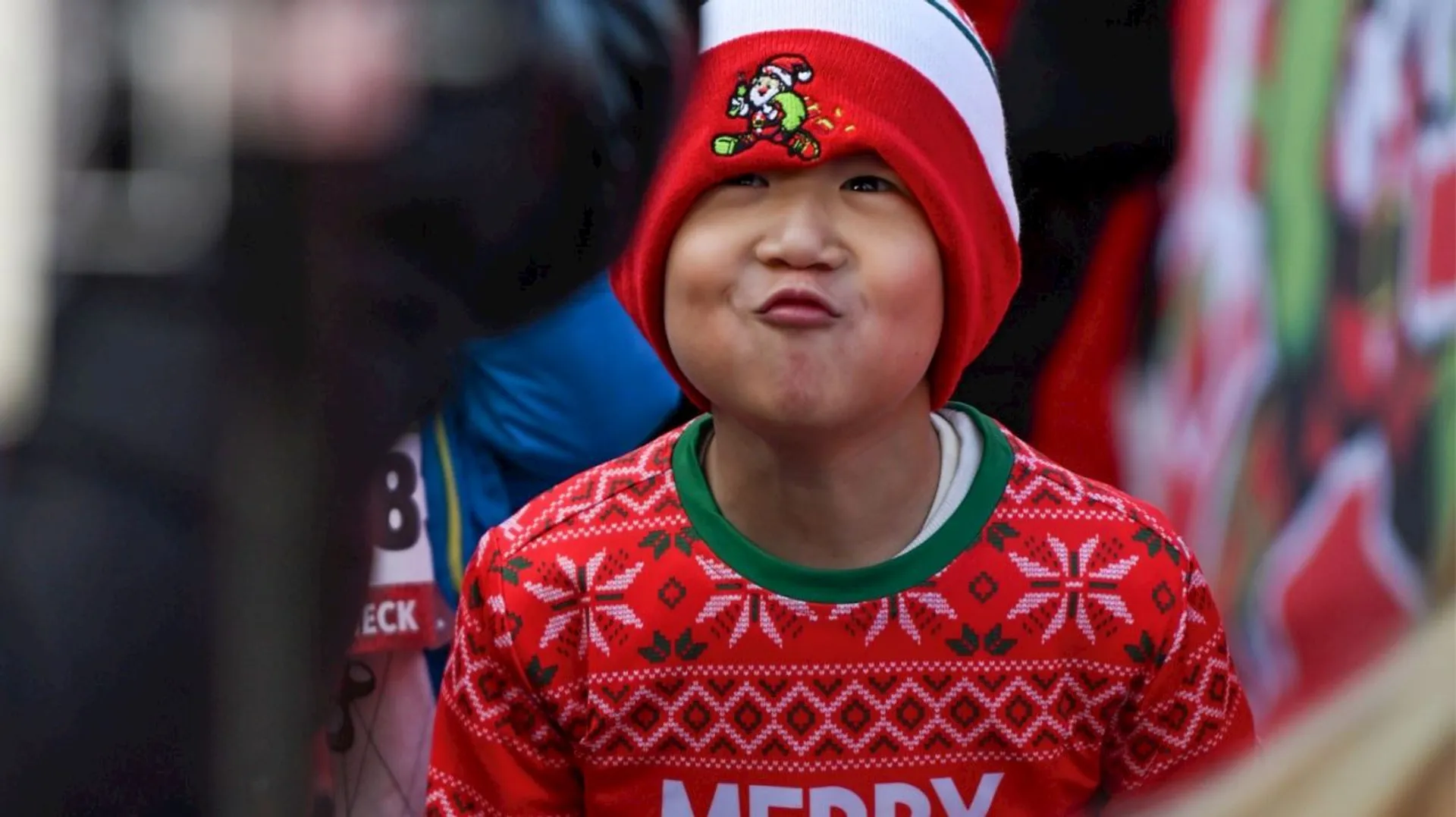 This image shows a child wearing a red sweater with snowflake patterns and the word "MERRY" on it. The child is also wearing a red and white hat with a small design on the front.