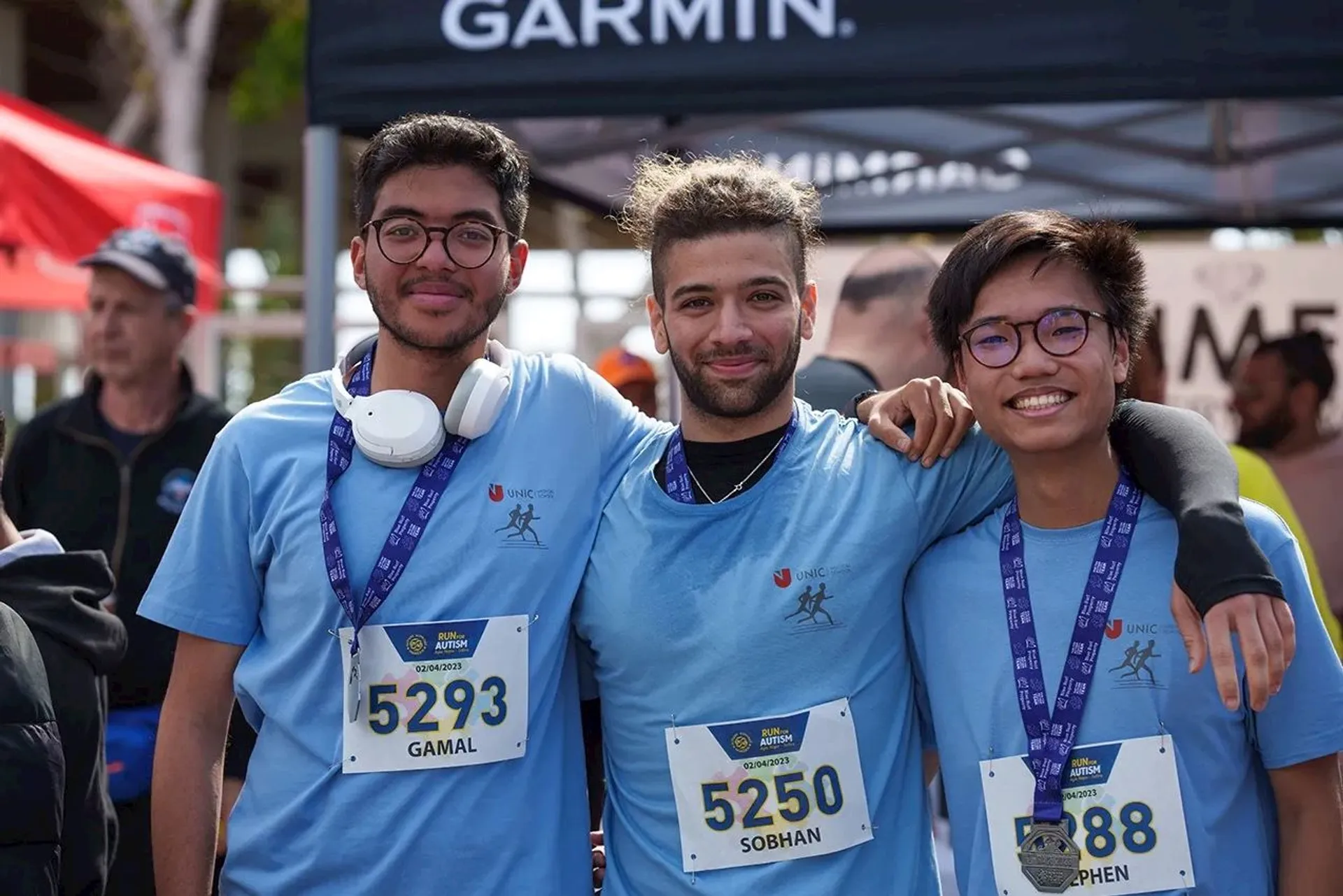 The image shows three people wearing light blue shirts and race numbers, standing closely together. They appear to be at a running event, as indicated by their race bibs and the background, which includes a Garmin tent. All three are smiling, and they have medals around their necks, suggesting they recently completed a race.