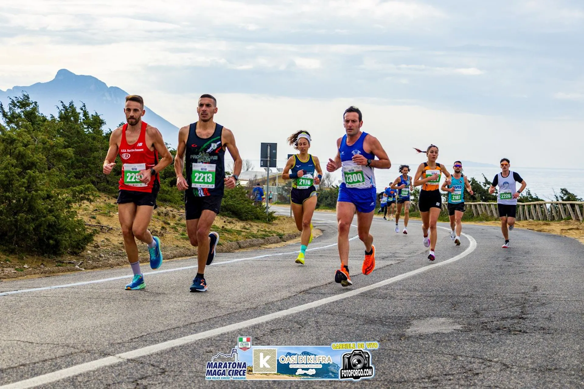 The image shows a group of runners participating in a road race. The event appears to be a marathon, as indicated by the banner at the bottom of the image. The road is beside a scenic coastline with mountains in the background. The participants have numbered bibs and are running on a winding road.