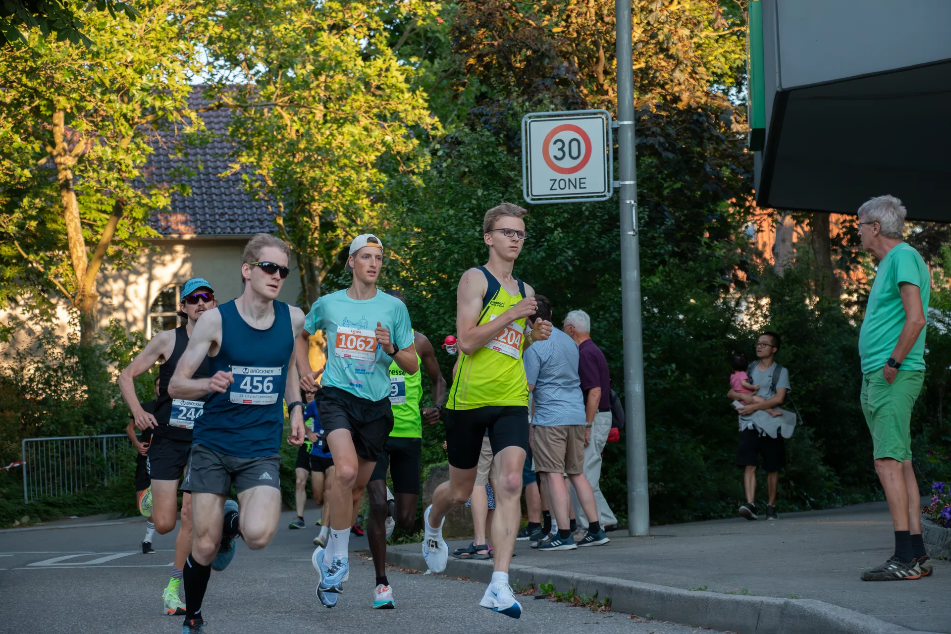 The image shows a group of runners participating in a road race. They are running past a street sign that indicates a 30 km/h speed limit zone, suggesting this might be a residential area or a place where reduced speed is required. There are also bystanders on the side, one of whom appears to be an official or volunteer associated with the race, as he is wearing a reflective vest. The trees and the lighting indicate that the race is taking place in the late afternoon or early evening. The runners are wearing bib numbers, which are typically used in organized races to identify participants. The environment looks calm and the race seems to be going on smoothly.