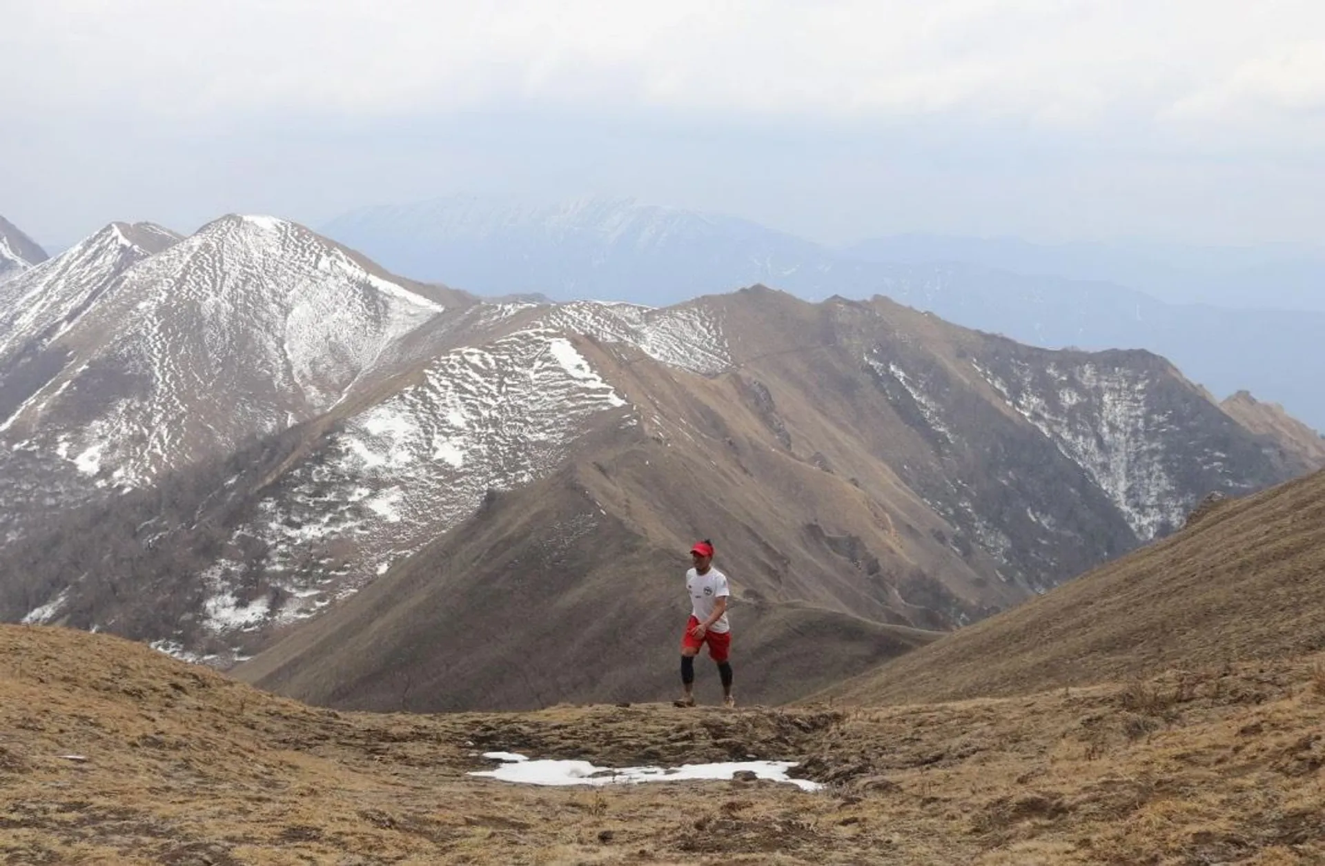 The image shows a person running or hiking on a mountainous terrain. The landscape features rugged, snow-dusted mountains in the background, with patches of snow still visible on some parts of the mountain slopes. The person is wearing athletic gear, including a red cap. The sky is cloudy, contributing to a somewhat overcast atmosphere.