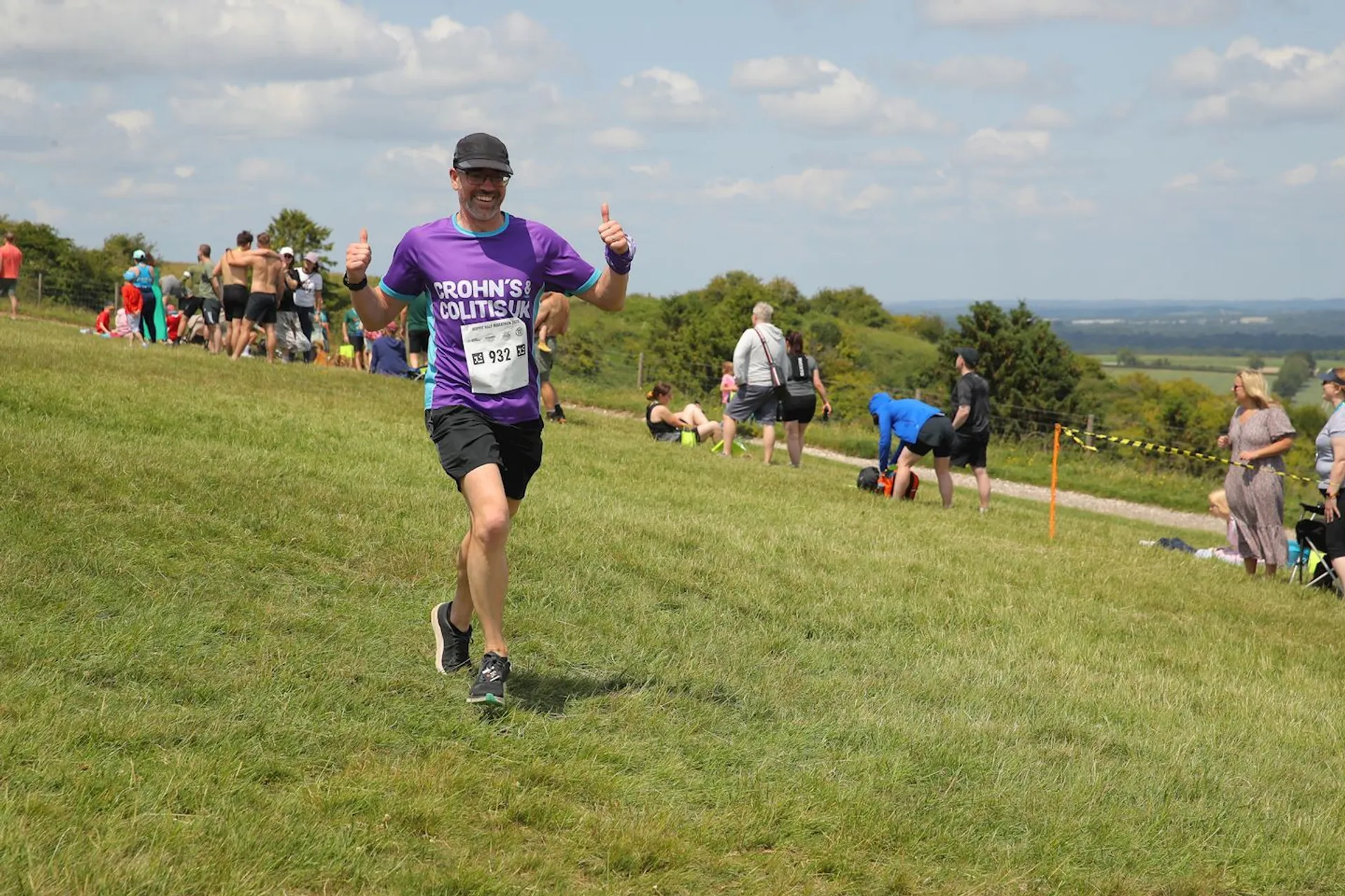 The image shows a person running outdoors in a grassy area during an event. The runner is wearing a purple T-shirt with a printed message, black shorts, and a cap, and is giving thumbs-up signs. Other people are nearby, some sitting on the grass, indicating it might be a race or gathering.