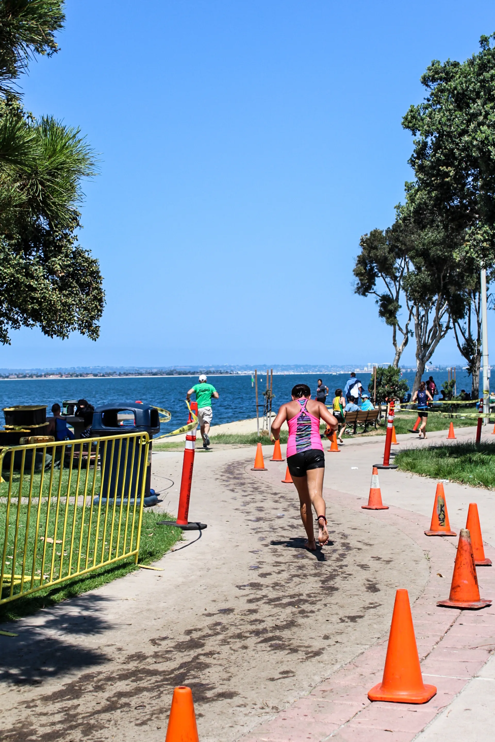 The image shows an outdoor setting with individuals participating in what appears to be a running event or race. There is a path lined with orange traffic cones to guide the runners. The path is bordered by greenery and trees, and in the background, there is a body of water, which could be a lake or sea. The sky is clear and blue, suggesting it's a sunny day. There are a few spectators or possibly fellow participants on the sides, and beyond the water, the horizon is visible. The setting looks like a park or a recreational area that is near a waterfront.