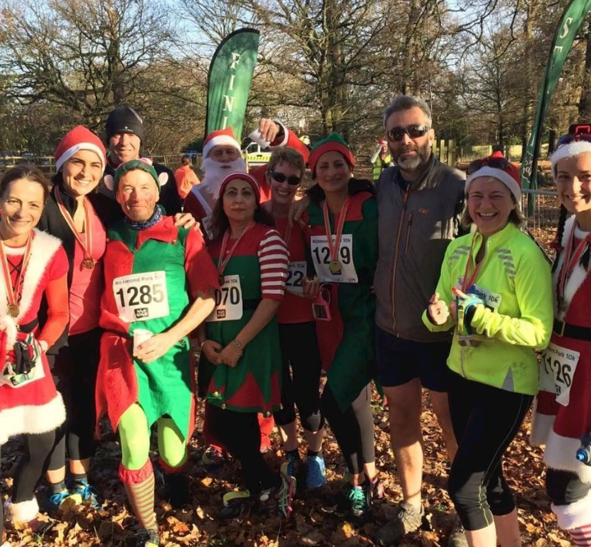 This image shows a group of people dressed in festive costumes, likely participating in a themed run or event. I see several people wearing Santa hats, and a few are dressed as elves with green outfits. The numbers pinned to their clothing suggest that they are participants in a race, possibly a fun run or charity event that is themed around Christmas. There are also inflatable structures that could be part of the race start or finish line. Everyone seems to be in high spirits and enjoying the occasion.