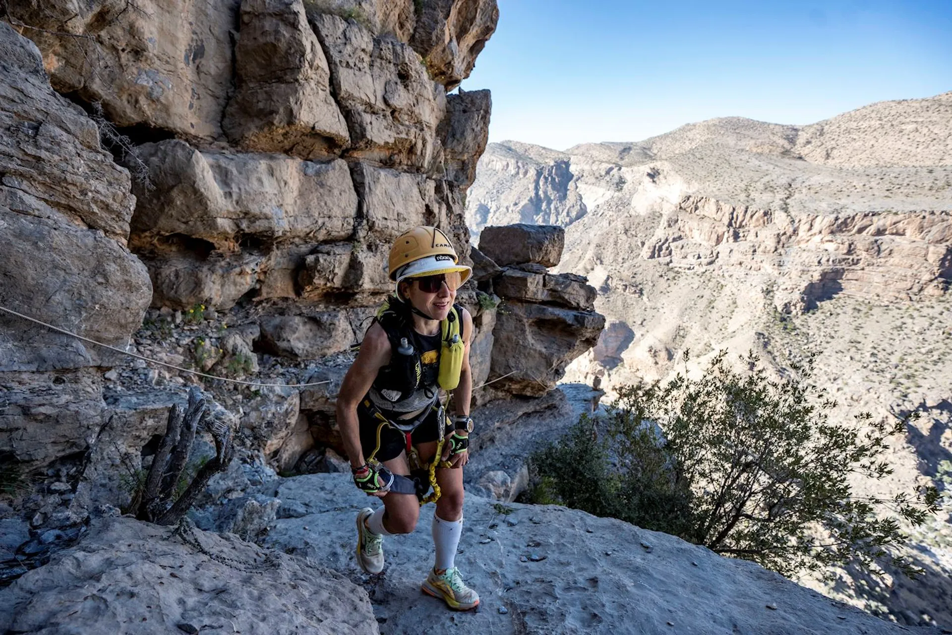 The image shows a person climbing or hiking along a rocky terrain, likely on a mountain trail. They are wearing a helmet, sunglasses, and are equipped with safety gear such as ropes and harnesses. The background features a dramatic landscape with rocky cliffs and a distant view of a canyon or mountainous area. The person appears to be engaged in a challenging outdoor adventure activity.