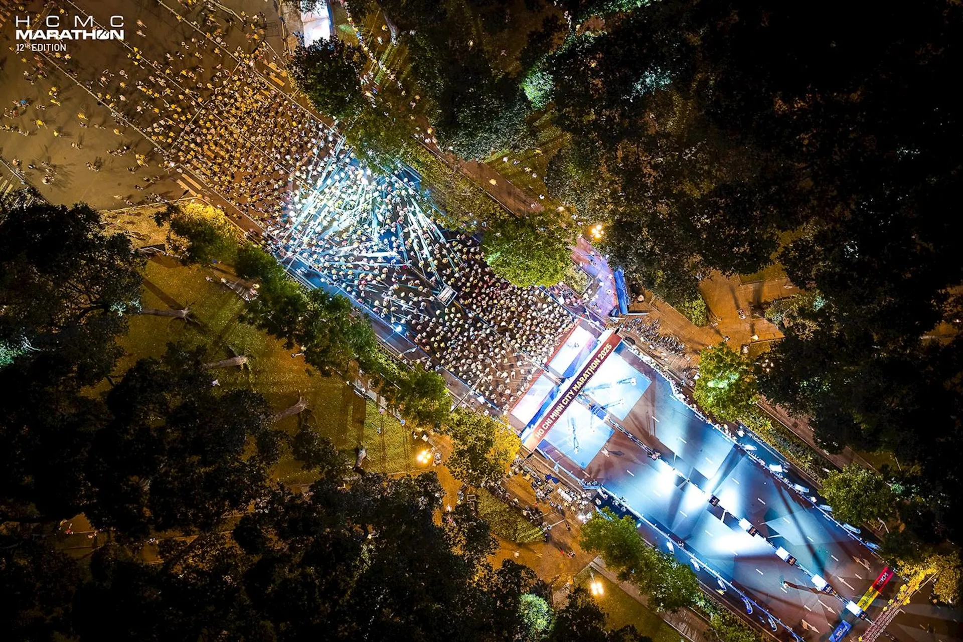 This is an aerial view of a marathon event. It shows a large group of people gathered at a starting line in a well-lit area, surrounded by trees. There are also several tents and structures, possibly for event support or spectators. The scene appears to be taken at night or early morning due to the use of artificial lighting.