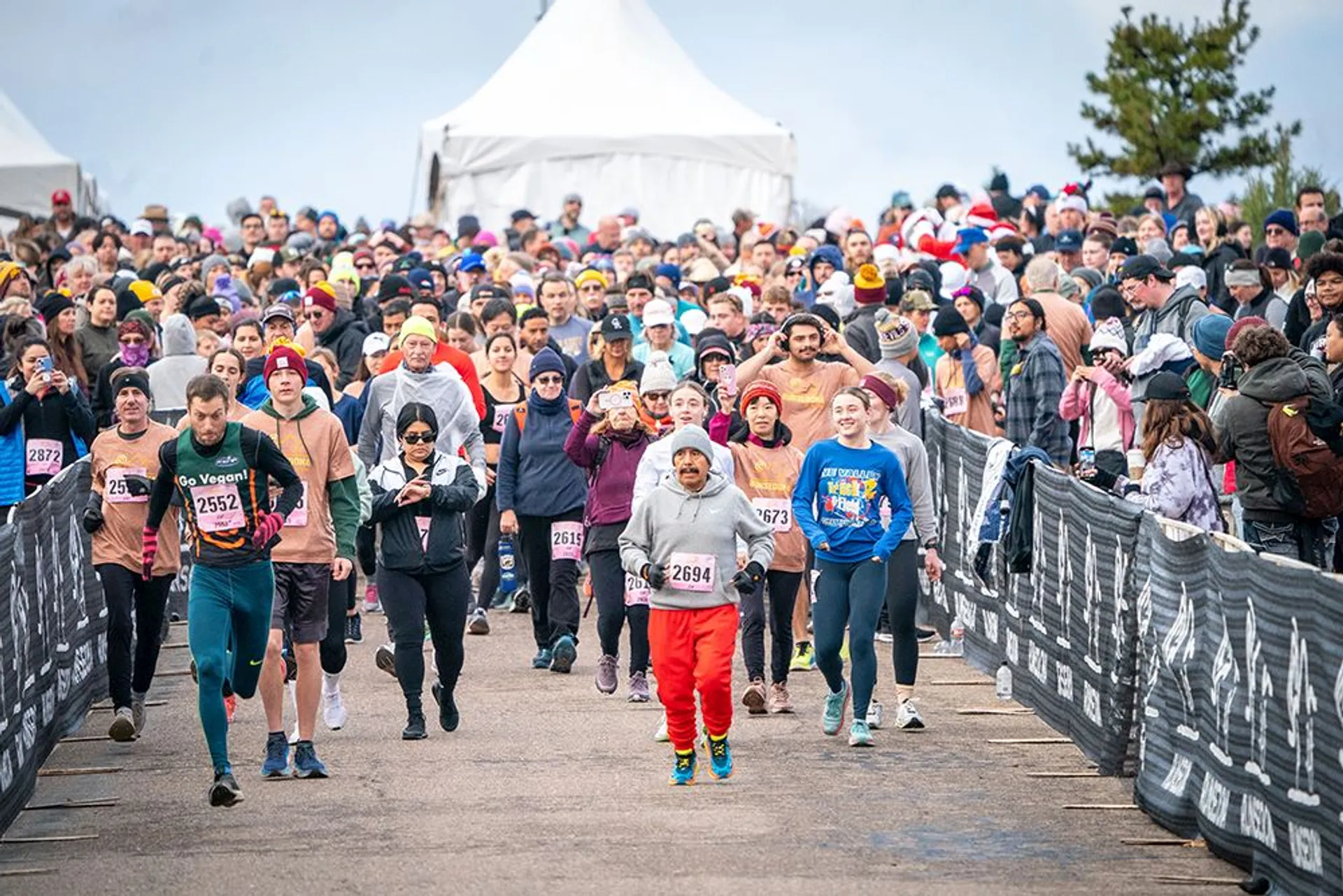 This image shows a large group of people participating in a running event. The runners are on a race course, surrounded by spectators behind barriers. Many participants are wearing numbered bibs and dressed in athletic wear. There are tents and trees in the background, and the event appears to be well-attended and lively.