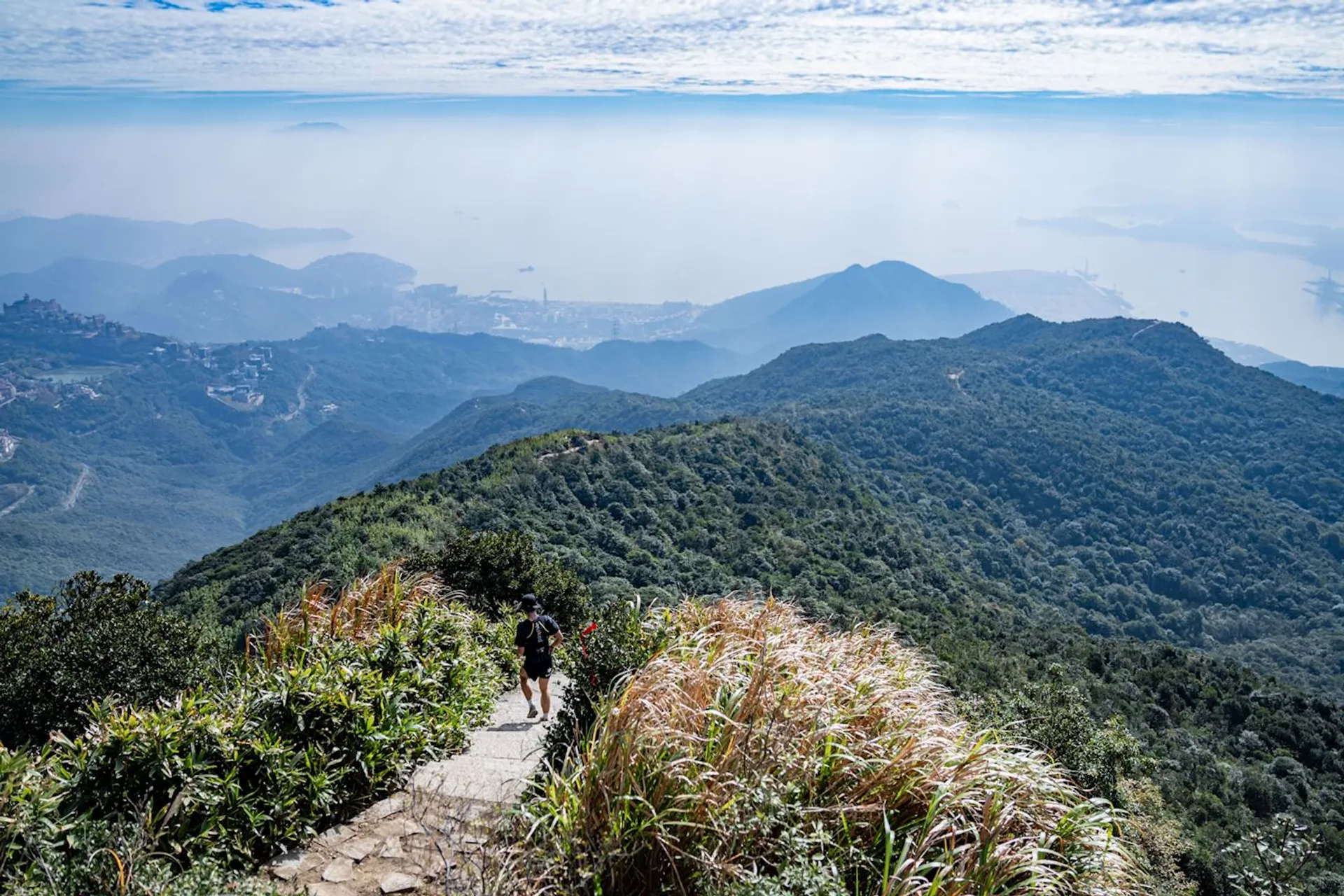 The image shows a scenic landscape from a high vantage point. A person is hiking on a trail that winds down the mountainous terrain. The vegetation appears lush with a mix of shrubs and grasses. In the distance, below the horizon of a seemingly hazy or cloudy sky, there's a panoramic view that suggests a coastal area, with lands stretching out towards a large body of water. The overall scene is one of natural beauty and suggests an environment that's ideal for outdoor activities like hiking. The alignment of the land and water indicates that this might be a high altitude viewpoint overlooking a coastline or a valley.