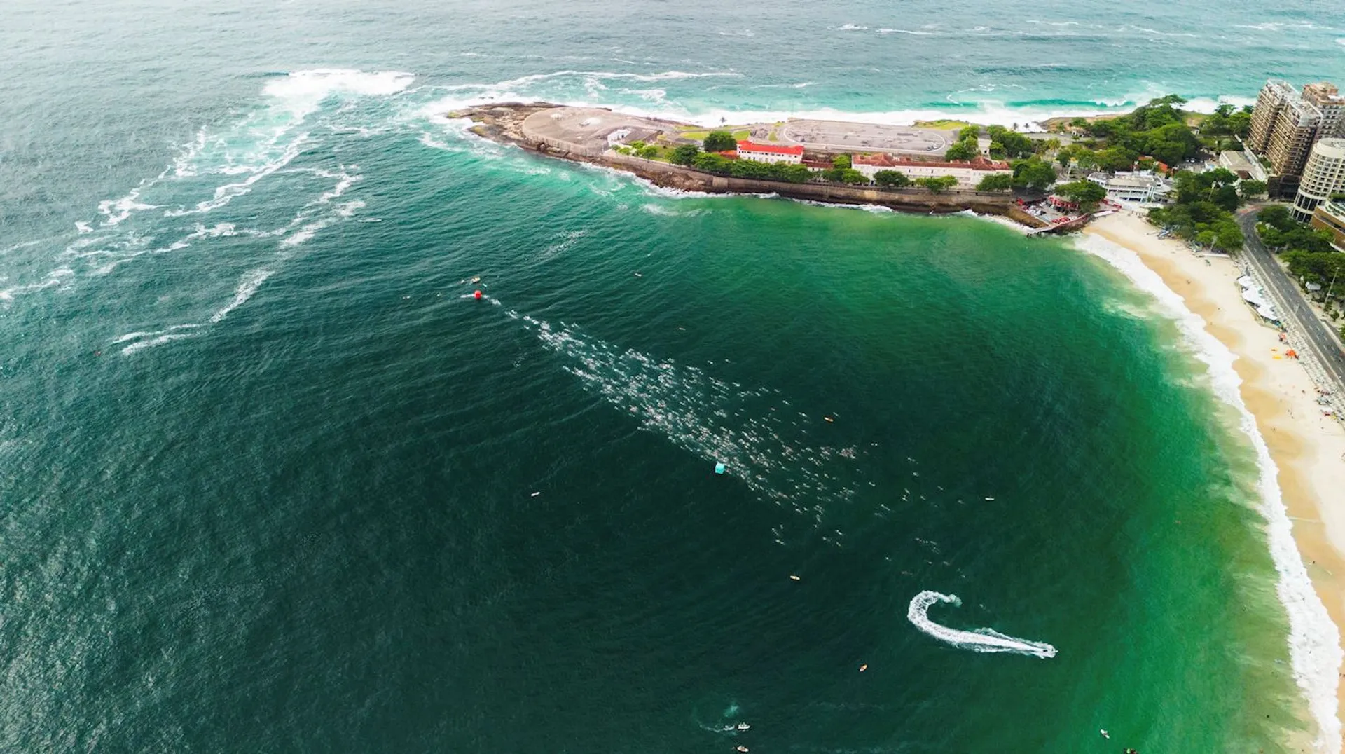 This image shows an aerial view of a coastal area with a beach. There is a large body of water with visible waves and a shoreline featuring sandy beaches. Near the shore, you can see people swimming or surfing in the ocean. There are land areas with vegetation and buildings, possibly a park or recreational area. The image captures a natural and recreational coastal scene.