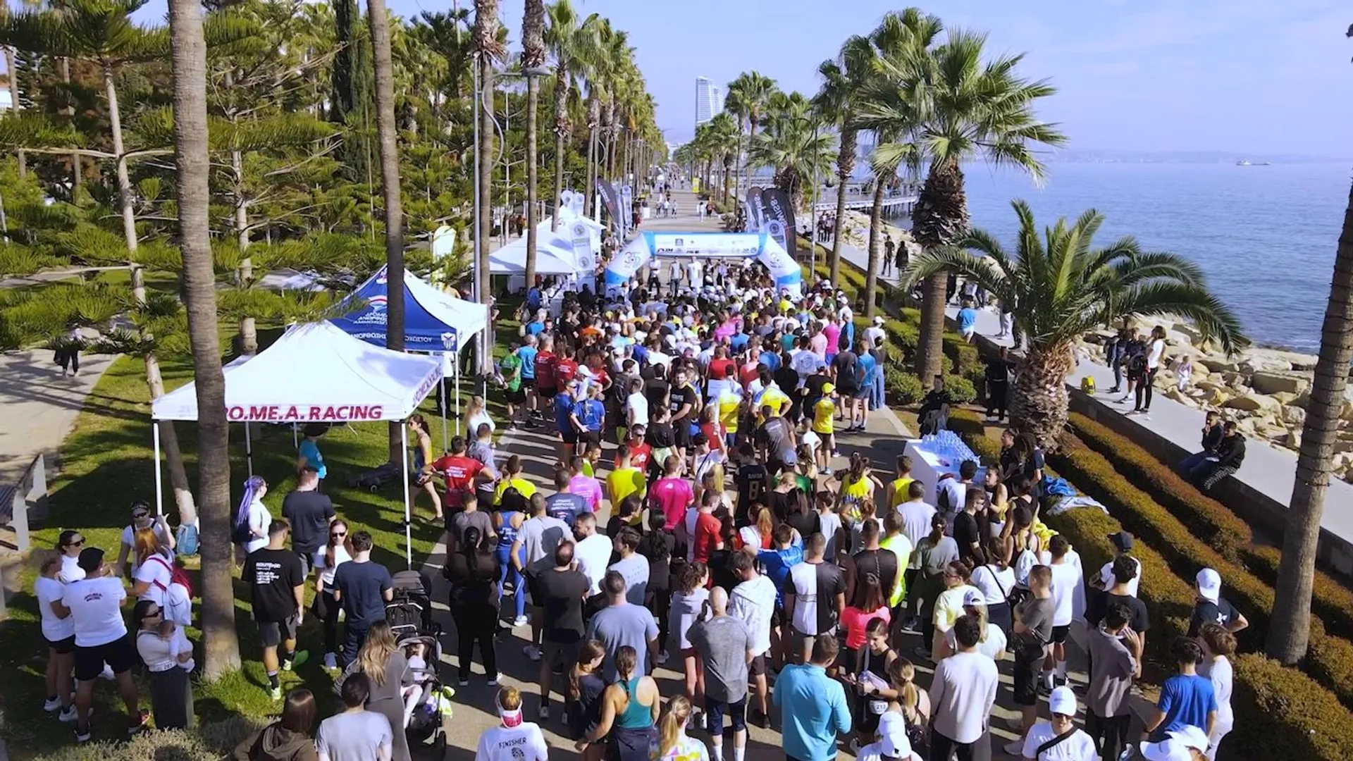 The image shows a large group of people gathered in what appears to be an outdoor event, possibly a race or a public gathering such as a marathon. The participants are in a waterfront area lined with palm trees, suggesting a coastal or beachside location. Tents and banners are set up, which may indicate sponsors or stations for the event. Some of the individuals are wearing running attire, supporting the idea that this might be a racing event. The environment looks sunny and clear, conducive to an outdoor activity. There are also views of the ocean and the horizon in the distance, emphasizing the scenic location of the event.