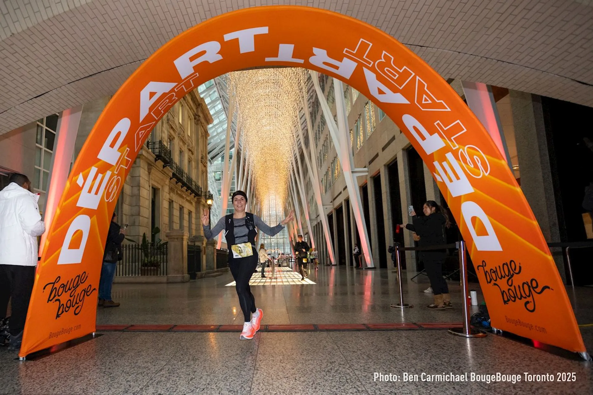 The image shows a person running through an archway that is marked with "DÉPART" and "START". The event seems to be organized by "BougeBouge" in Toronto, as indicated by the text on the arch and the photo credit. The arch is set up indoors, and there's a decorative light installation in the background.