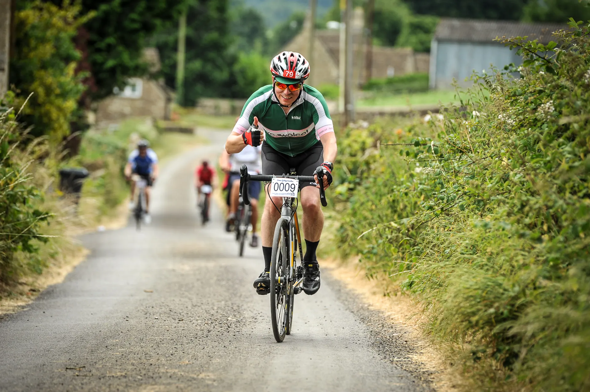 The image depicts a group of cyclists participating in a bicycling event or race. In the foreground, there's a cyclist wearing a white helmet, sunglasses, and a cycling jersey with green accents, and a number visible with "0009". The cyclist is focused and seems to be pedaling with effort. The background shows other participants following and a rural or countryside setting with foliage on either side of a narrow road. It looks like a day of active outdoor sports, and all individuals seem engaged in the activity.