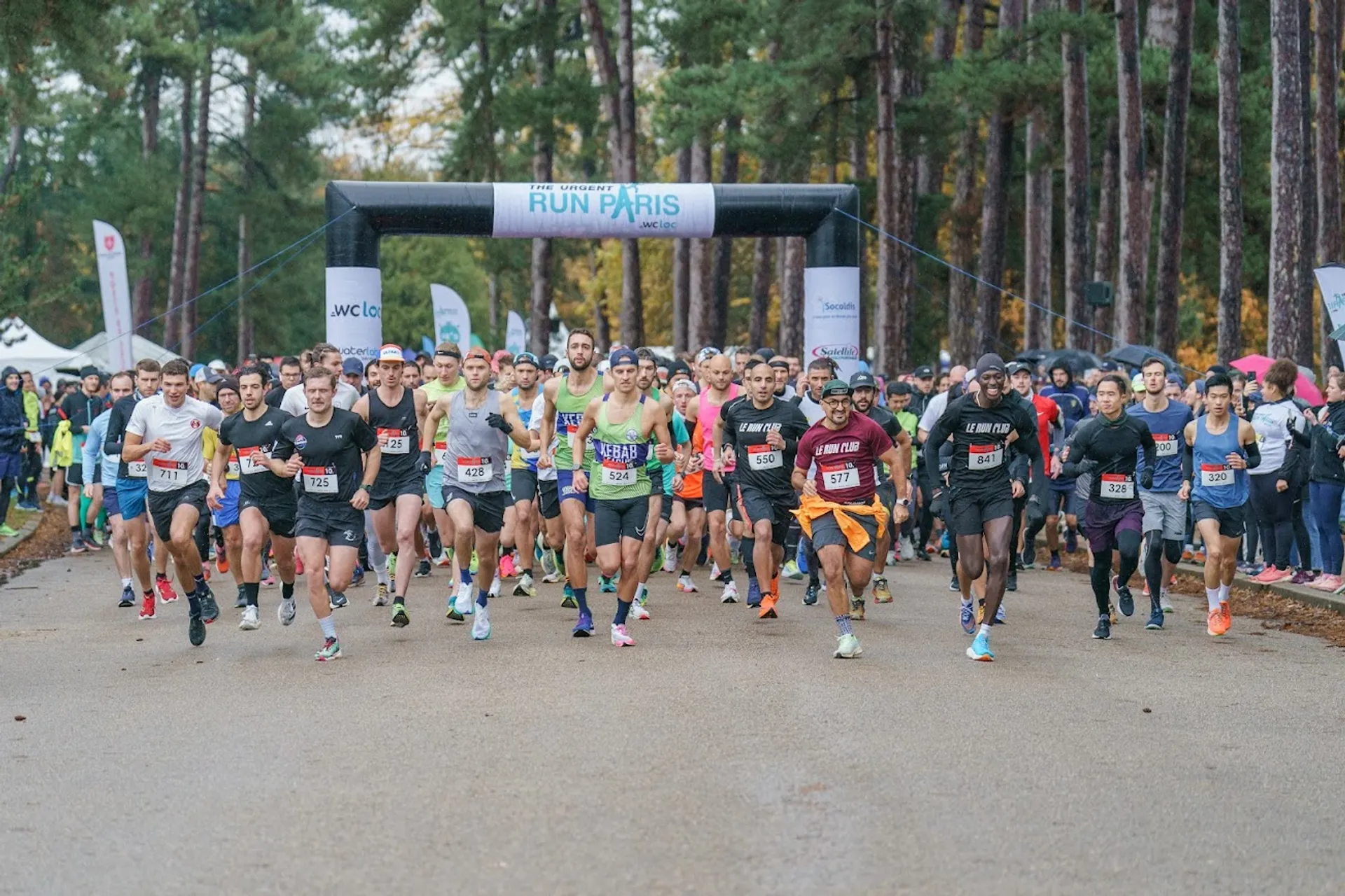 This image depicts a group of runners at the start of a race. The participants are just crossing the starting line, which is marked by a large banner reading 'RUN PARIS'. They are in a variety of running attire, suggesting that this is a diverse group of individuals with different levels of experience and preparedness for the race. The setting appears to be a park or a wooded area, and it seems to be an overcast day, based on the lighting and the attire of the participants. On either side of the runners, there are barricades and several tents, which are likely associated with the event, possibly for registration, information, or as aid stations. The event seems well-organized, with a clear starting line and visible signage.