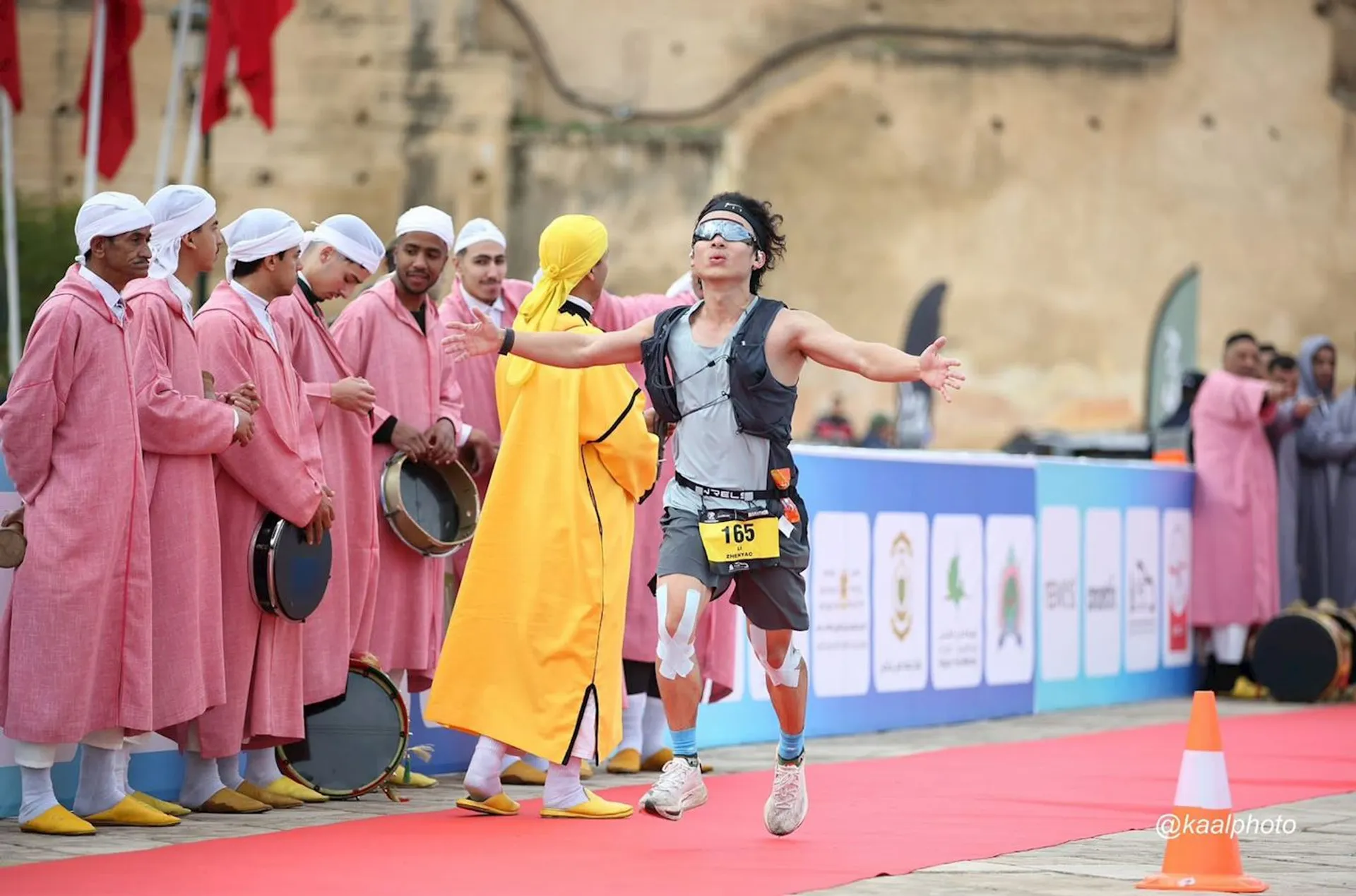 The image depicts a runner crossing a finish line during a race event. The runner is wearing a race bib and appears to be celebrating. In the background, there are people dressed in traditional attire, some of whom are playing musical instruments like drums. The setting includes a red carpet marking the finish line and banners with various logos and sponsors.
